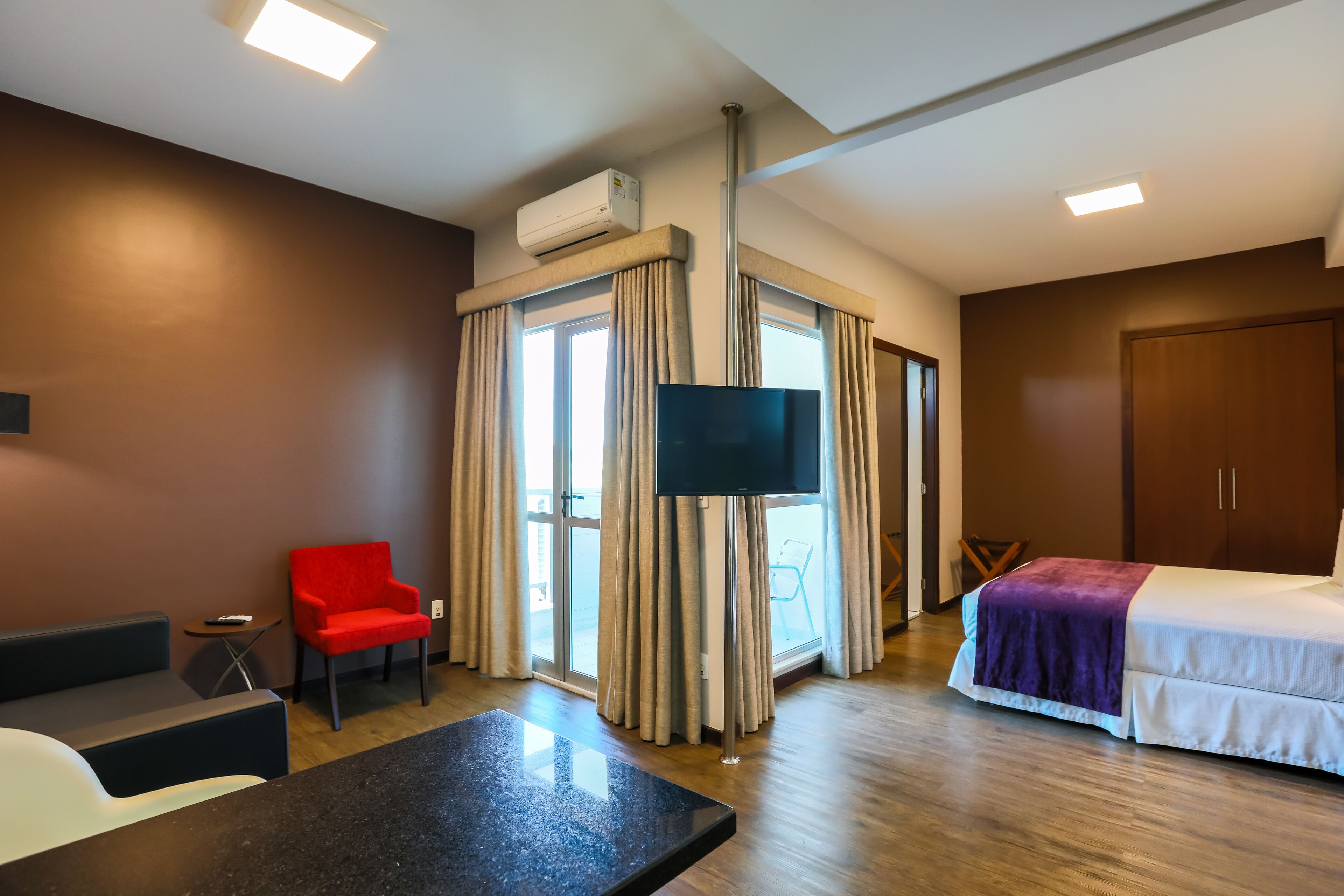 A hotel room with a bed, a TV, a red chair, a small table, and a window with curtains.