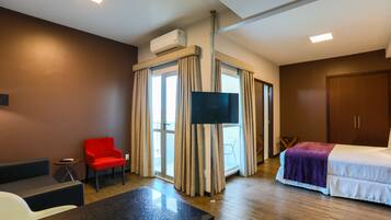 A hotel room with a bed, a TV, a red chair, a small table, and a window with curtains.