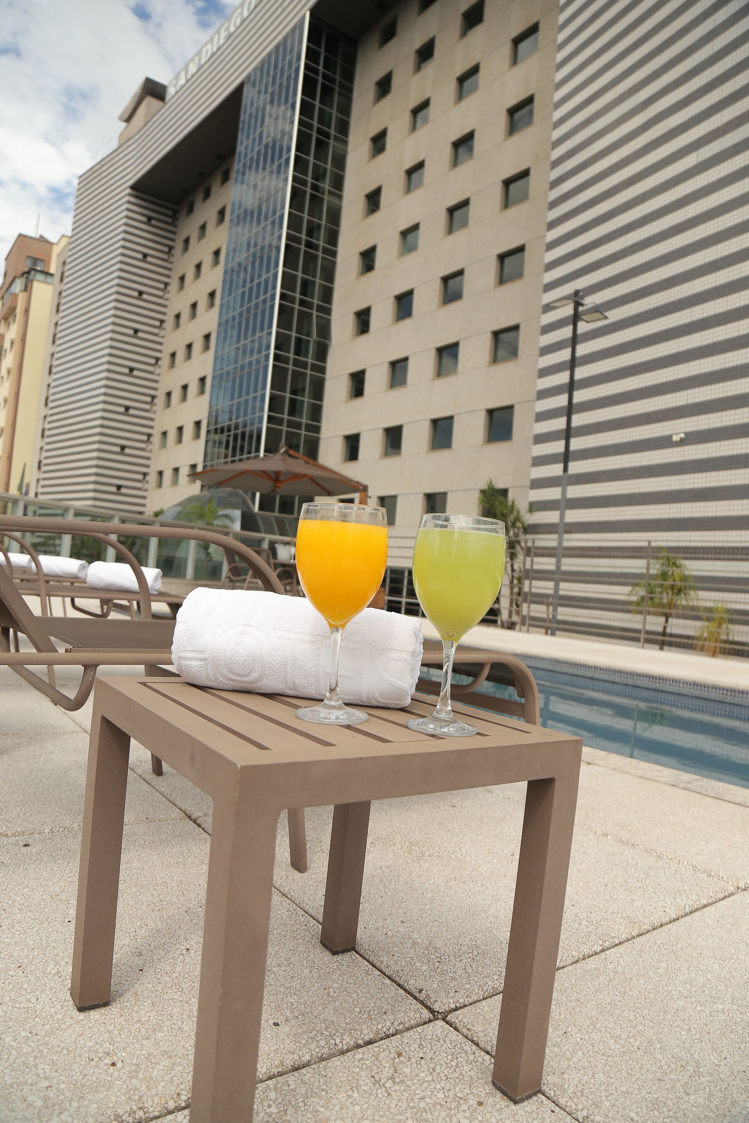 Mesa à beira da piscina com dois copos de sumo de laranja e um cocktail verde, toalhas brancas e um edifício moderno ao fundo.
