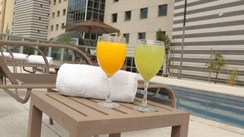Mesa à beira da piscina com dois copos de sumo de laranja e um cocktail verde, toalhas brancas e um edifício moderno ao fundo.