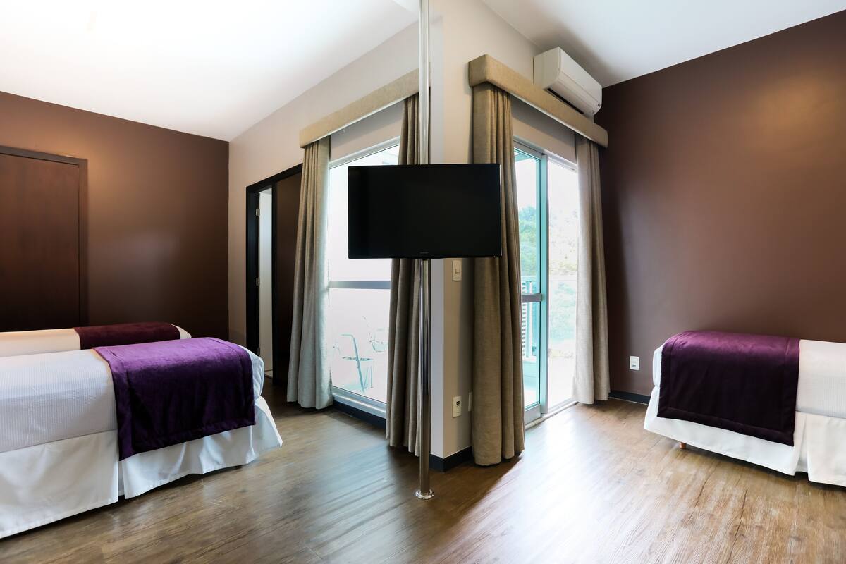 A hotel room with two beds, a flat-screen TV, a large window with curtains, and a wooden floor.