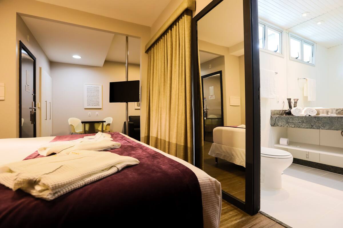 A hotel room with a bed, a desk, a chair, a television, and a bathroom with a toilet and sink.