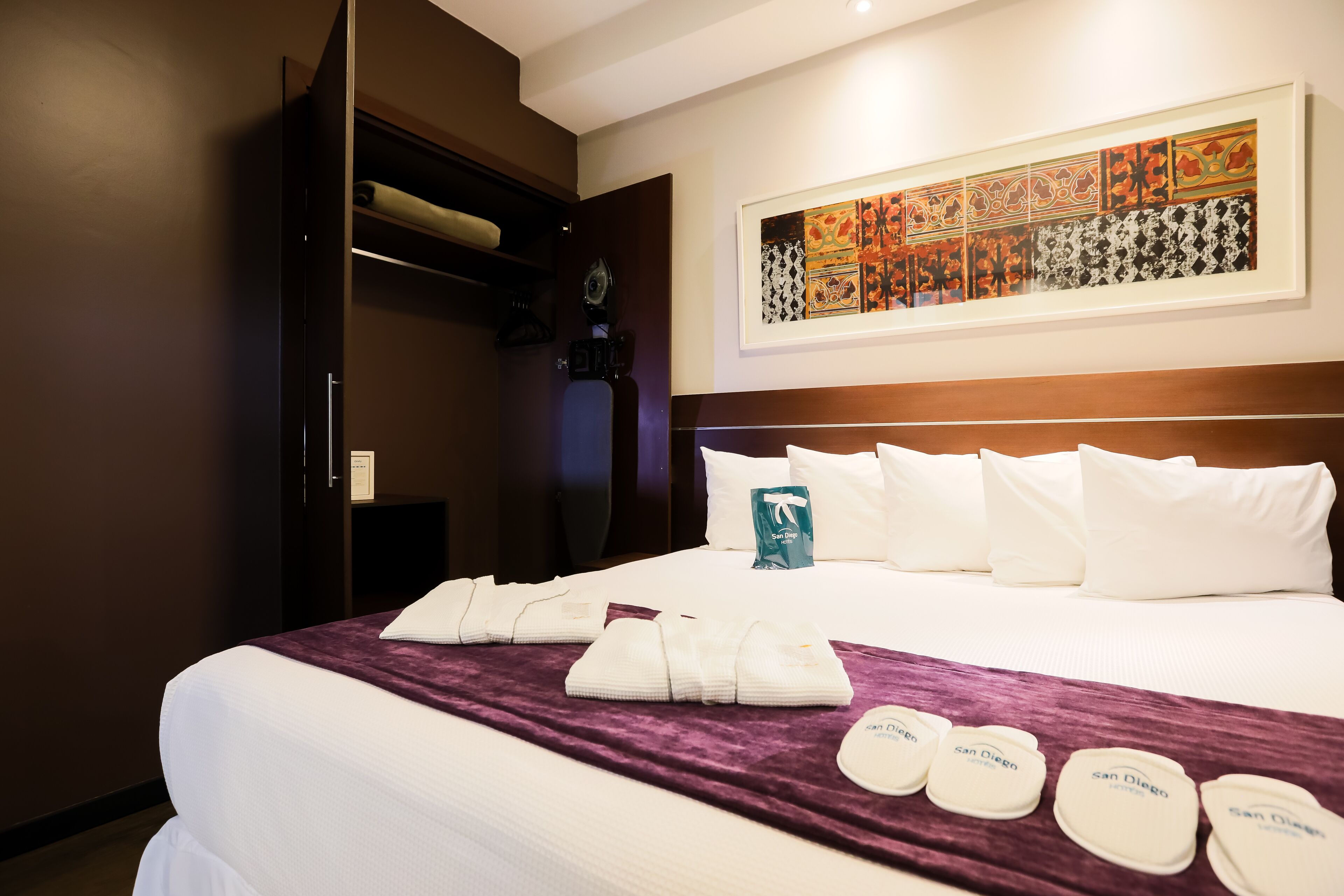 A hotel room with a neatly made bed, a large artwork on the wall, and a closet with a mirror.