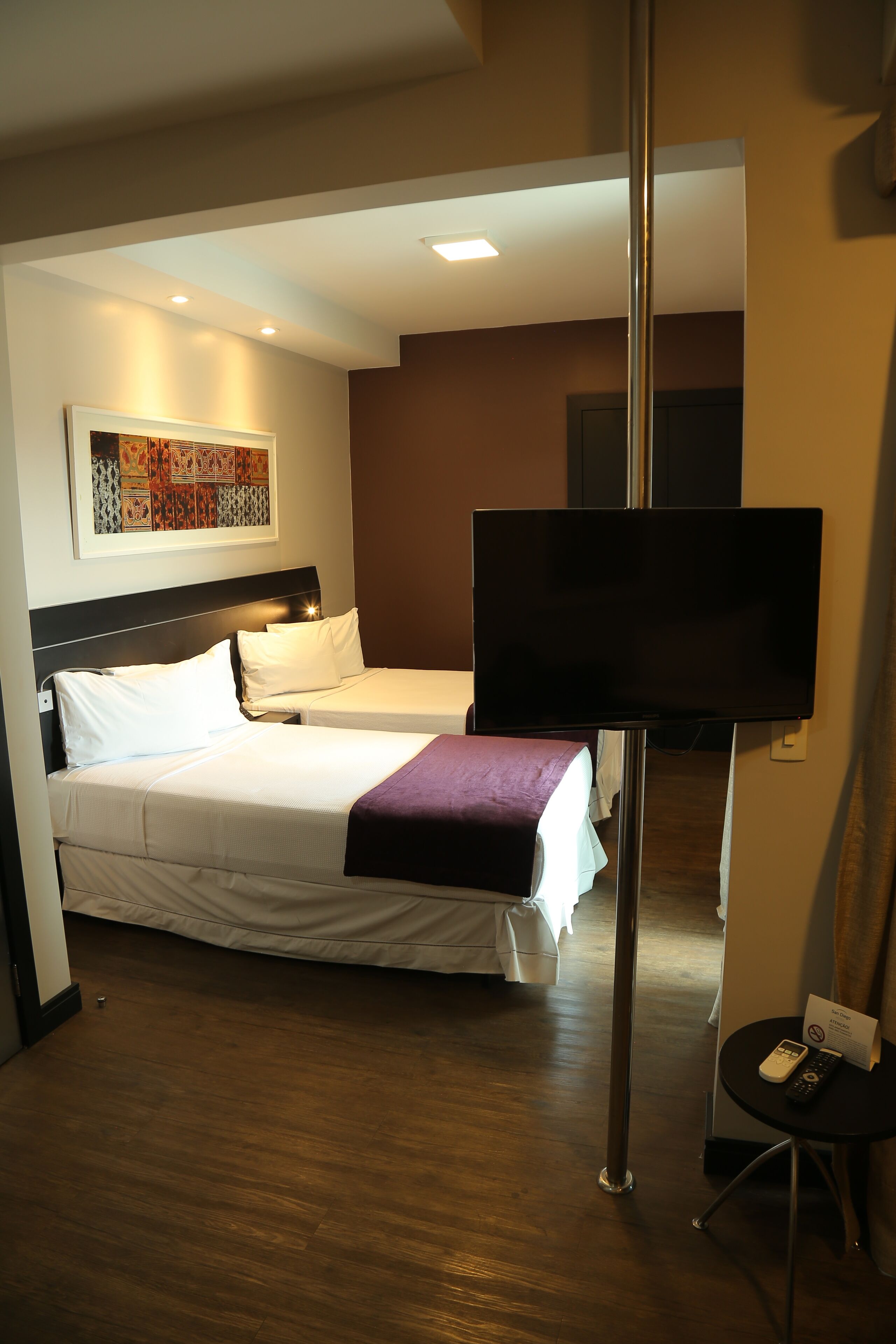 A hotel room with two beds, a flat-screen TV mounted on a pole, a small table with a remote control, and a framed artwork on the wall.