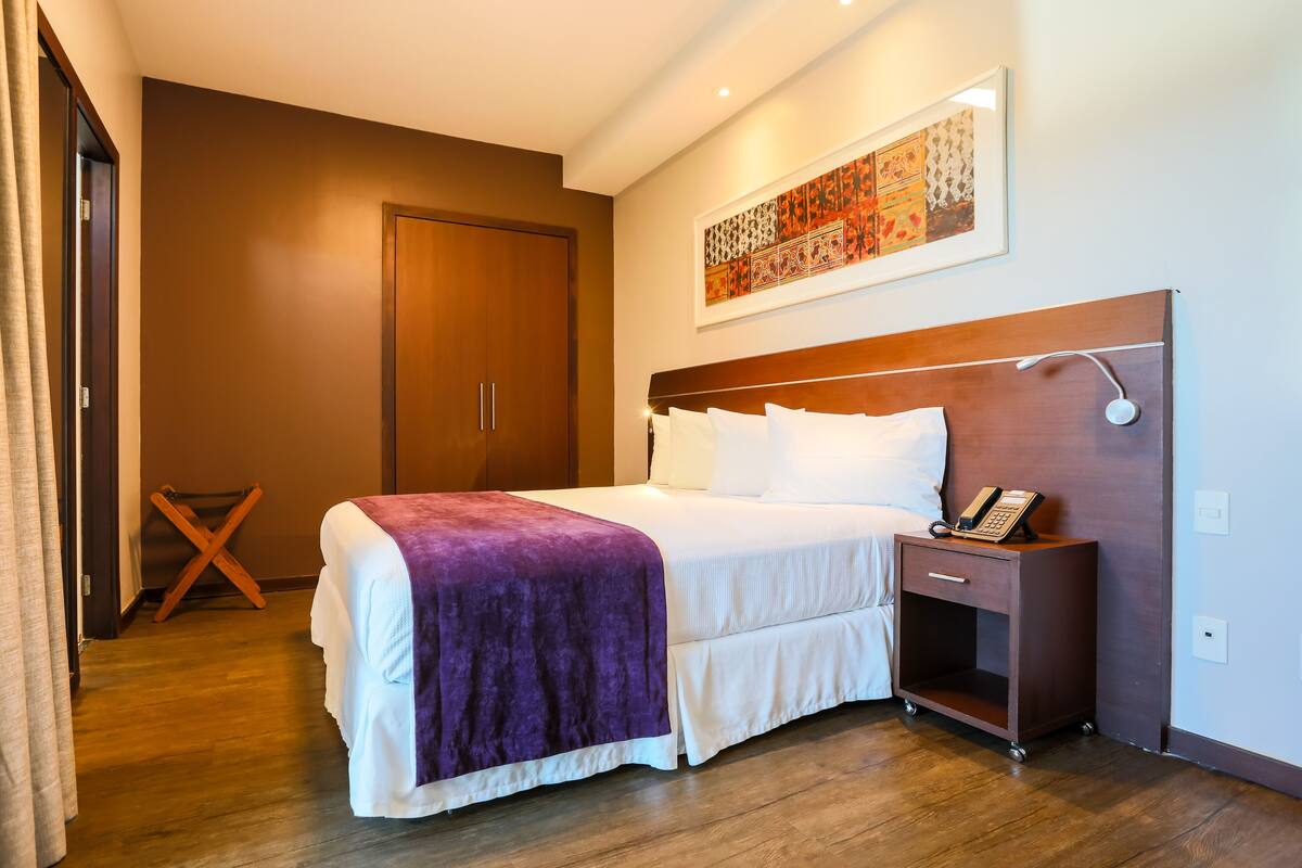 A hotel room with a large bed, a wooden headboard, a bedside table with a phone, and a wall-mounted artwork.