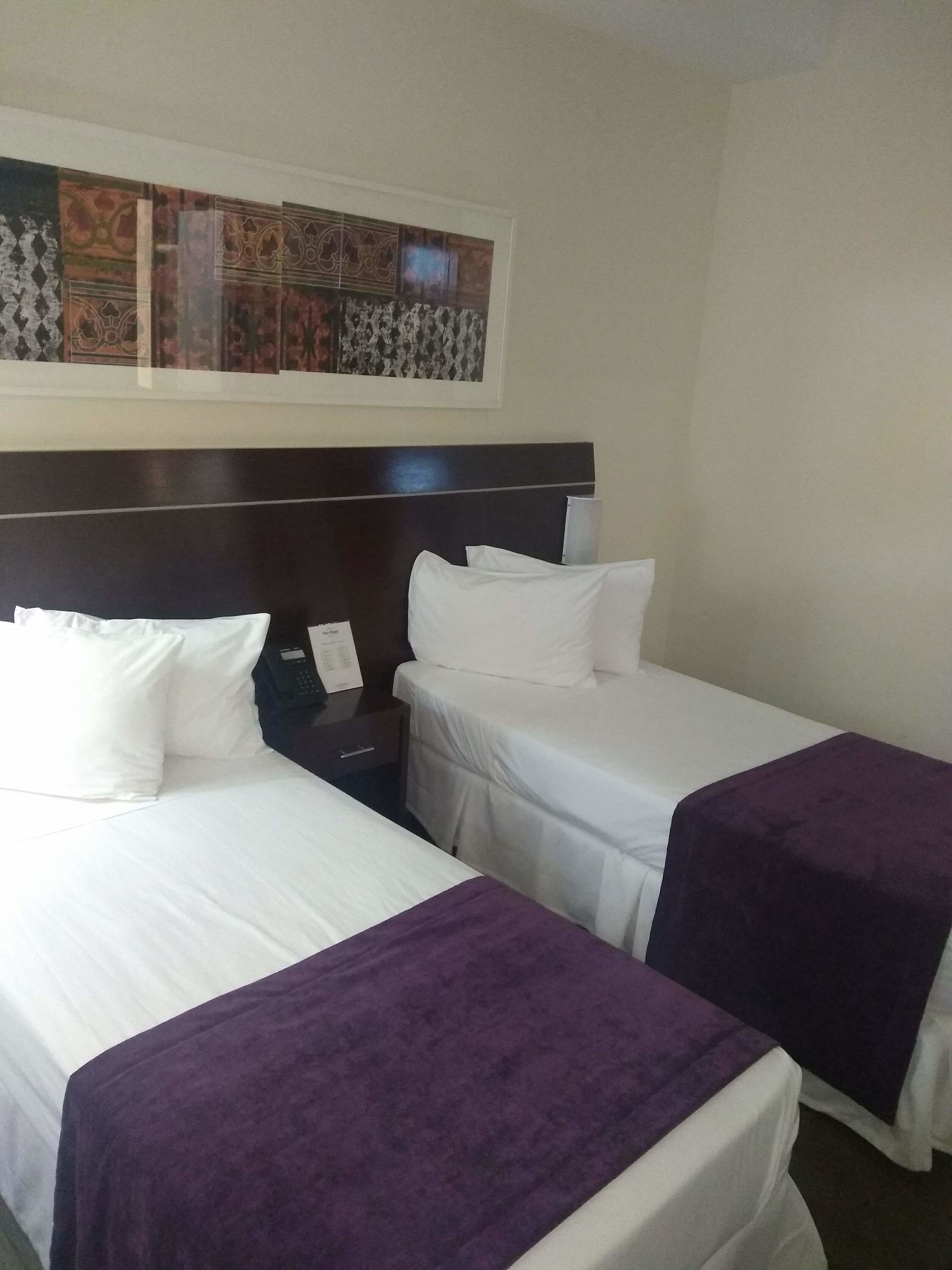 Una camera d'albergo con due letti, entrambi con lenzuola bianche e una coperta viola. Si nota un poggiatesta in legno scuro, un comodino con un telefono e un quadro incorniciato sopra i letti.