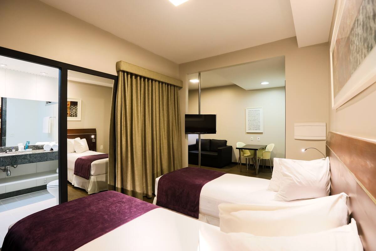 A hotel room with two beds, a desk, a chair, a TV, and a bathroom visible through a glass partition.