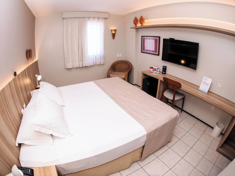 Una habitación de hotel con cama, escritorio, silla, televisión y ventana con cortinas.