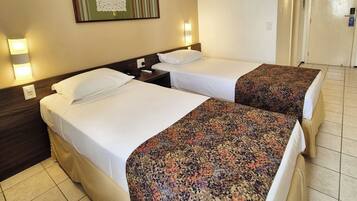 Habitación de hotel con dos camas, cada una con ropa blanca y una manta decorativa. Hay una obra de arte enmarcada sobre las camas y una lámpara de pared a la izquierda.