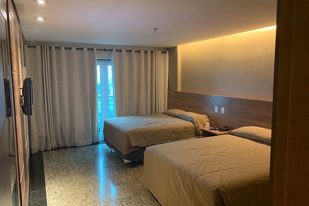 A hotel room with two beds, a wooden headboard, a flat-screen TV, and a view of the outdoors through a window.