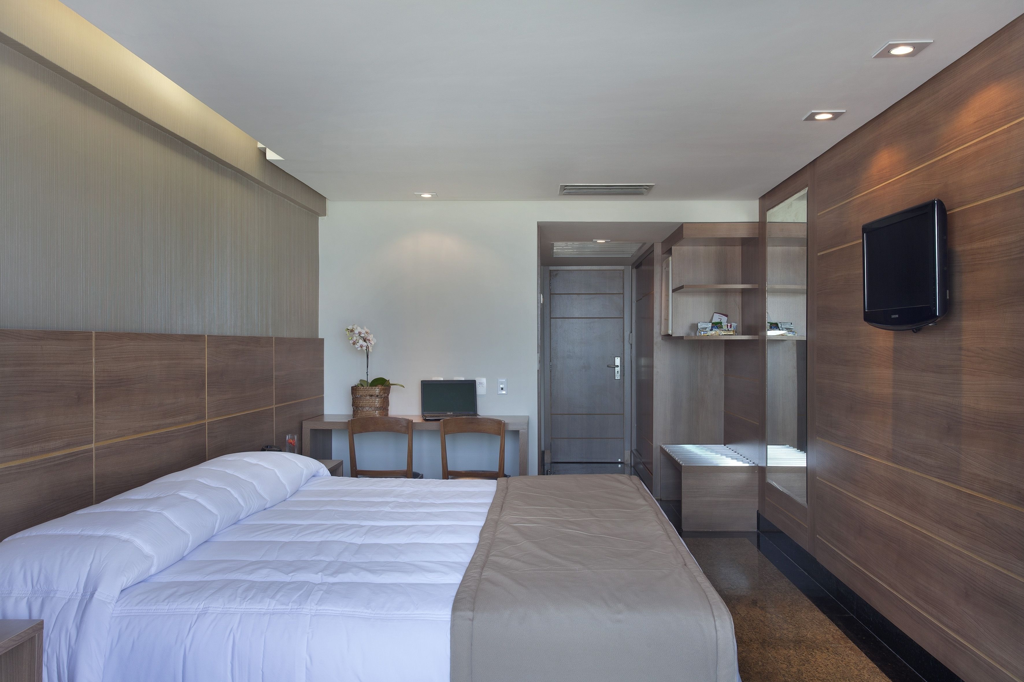 A modern hotel room with a large bed, a desk with a computer, a TV mounted on the wall, and a wardrobe.