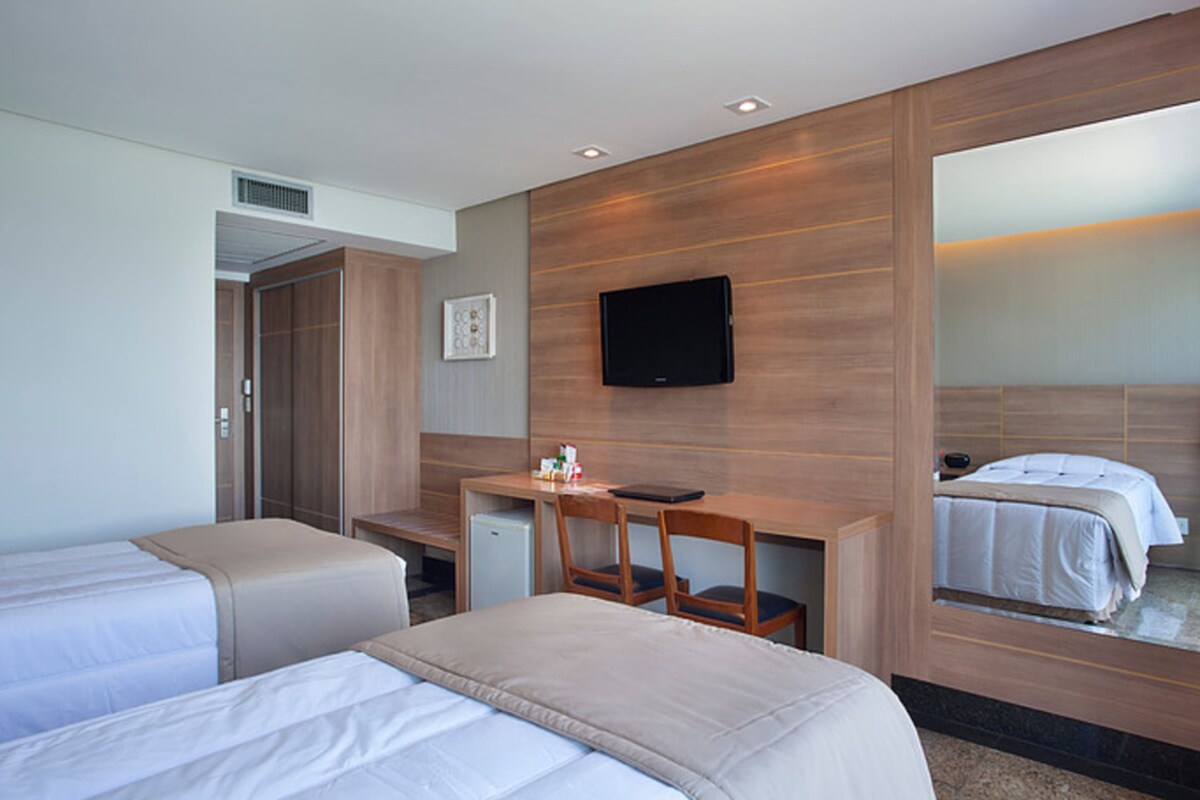 Ein Hotelzimmer mit zwei Betten, einem Schreibtisch, einem Sessel, einem Fernseher und einem Spiegel.