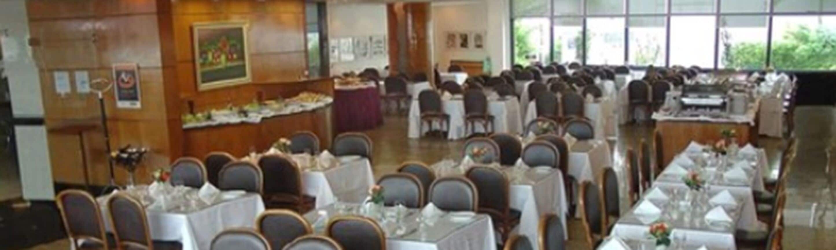 Salão de festas com mesas dispostas em fileiras para um evento formal, com teto alto e grandes janelas.