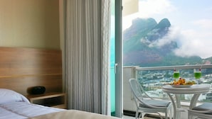 A hotel room with a bed, a bedside table with a radio, a balcony with a table, chairs, and a view of mountains and a city. - Royalty Barra Hotel (Rio de Janeiro)