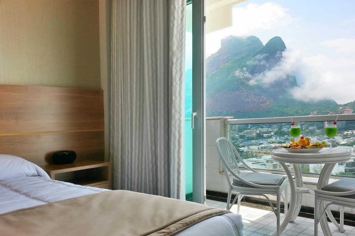 Une chambre d’hôtel avec un lit, une table de chevet avec une radio, un balcon avec une table, des chaises et une vue sur les montagnes et la ville.
