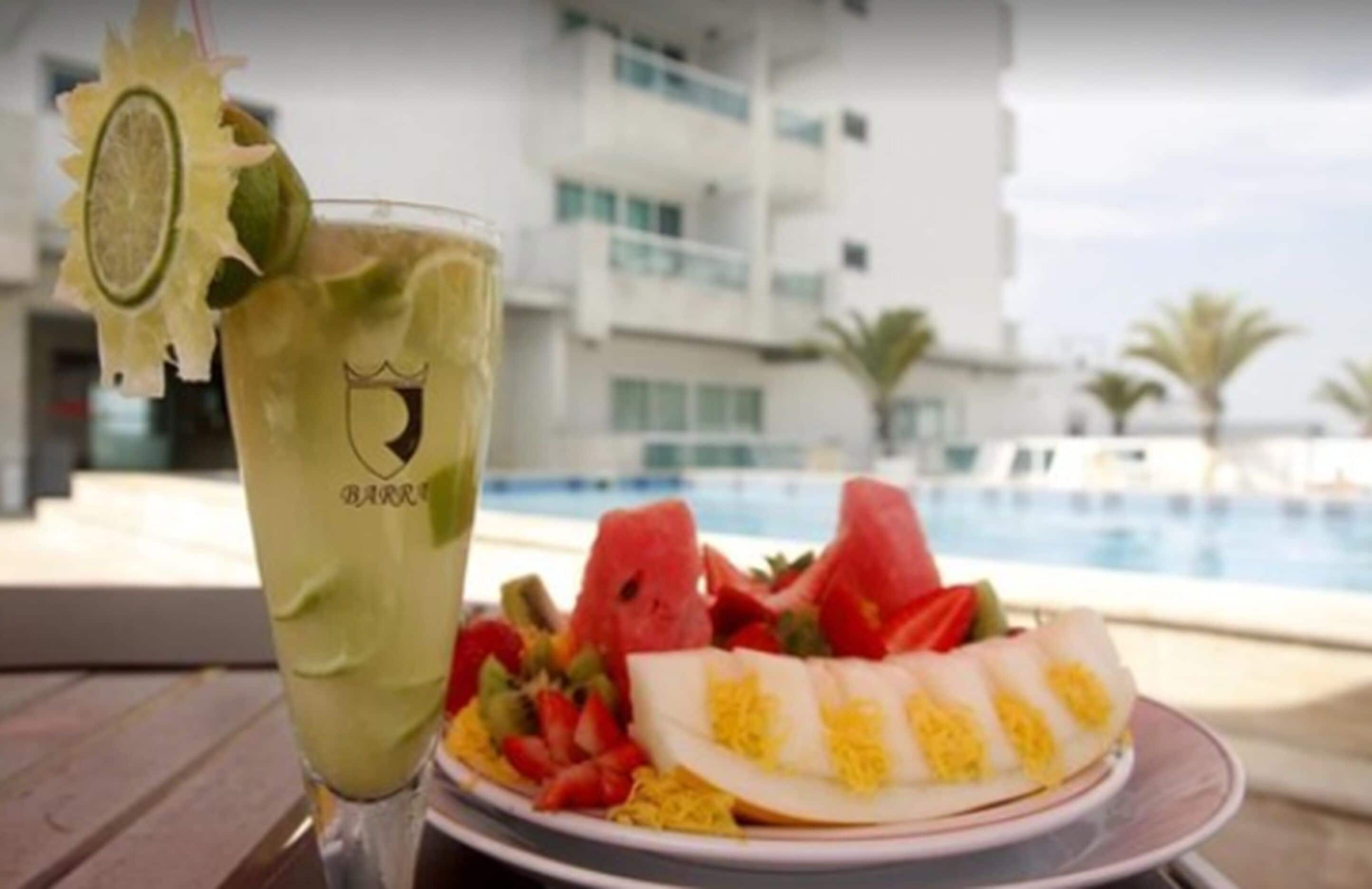 A drink with a lime garnish and a plate of fruit next to a poolside view of a building.