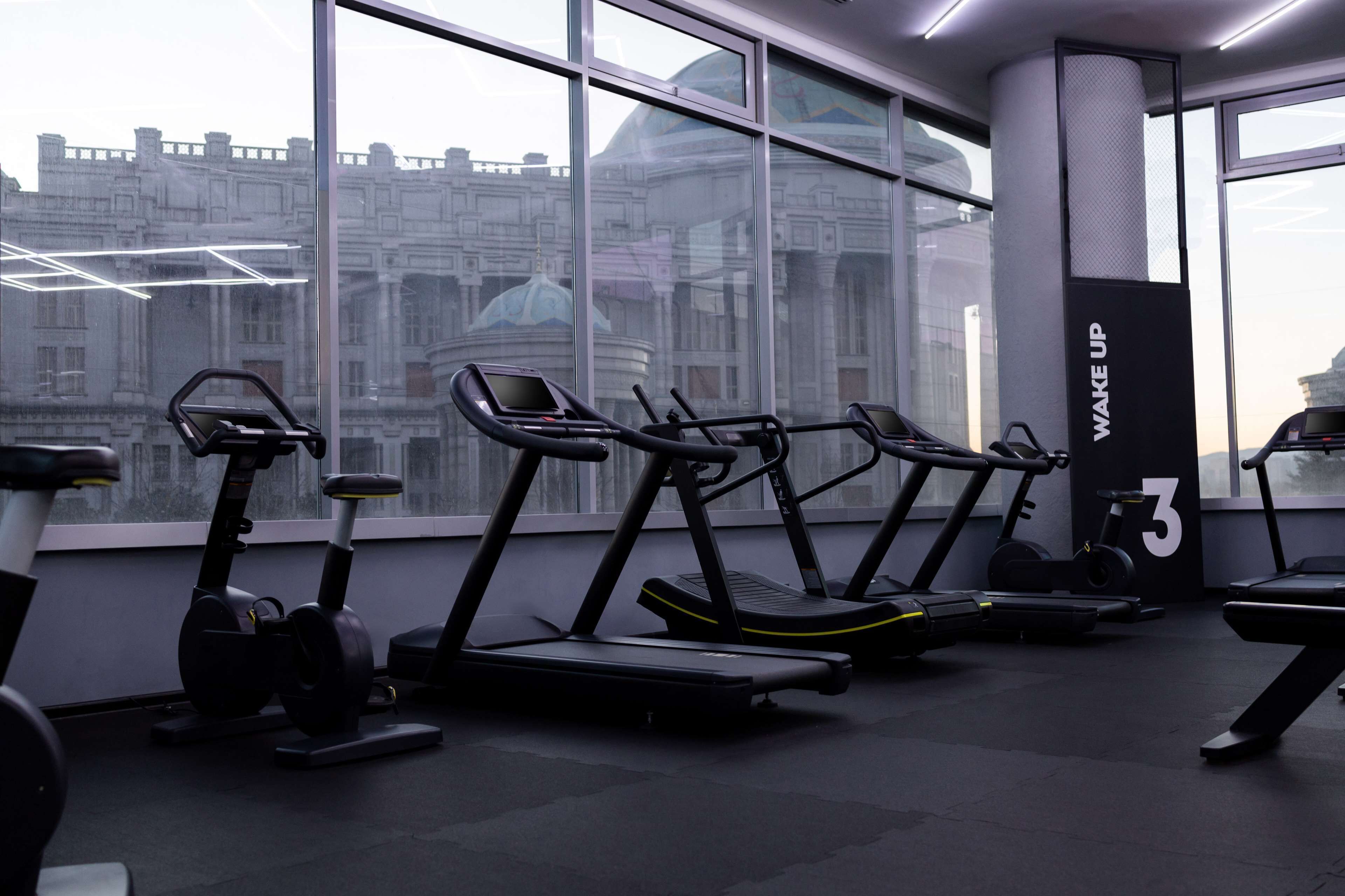 Une salle de sport équipée de tapis de course et de vélos d’appartement, de grandes fenêtres offrant une vue sur la ville et un panneau indiquant « Réveillez-vous ».