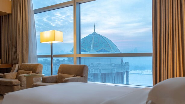 A hotel room with a large bed, two armchairs, a small table, and a floor lamp. A large window offers a view of a cityscape and a dome structure.
