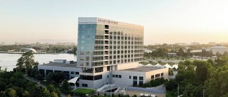 Un moderno hotel Hyatt Regency con fachada de vidrio, rodeado de vegetación y un río.