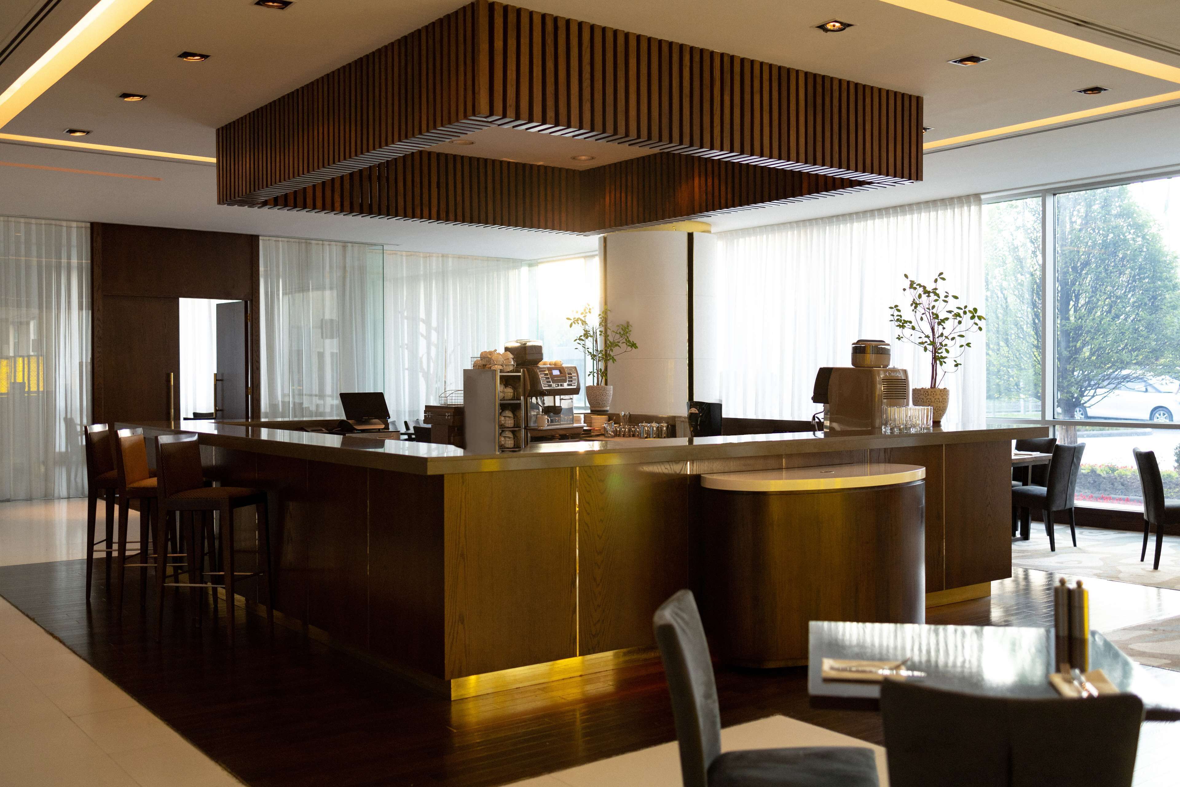 A modern hotel lobby with a central reception desk, seating area, and large windows offering a view of the outdoors.