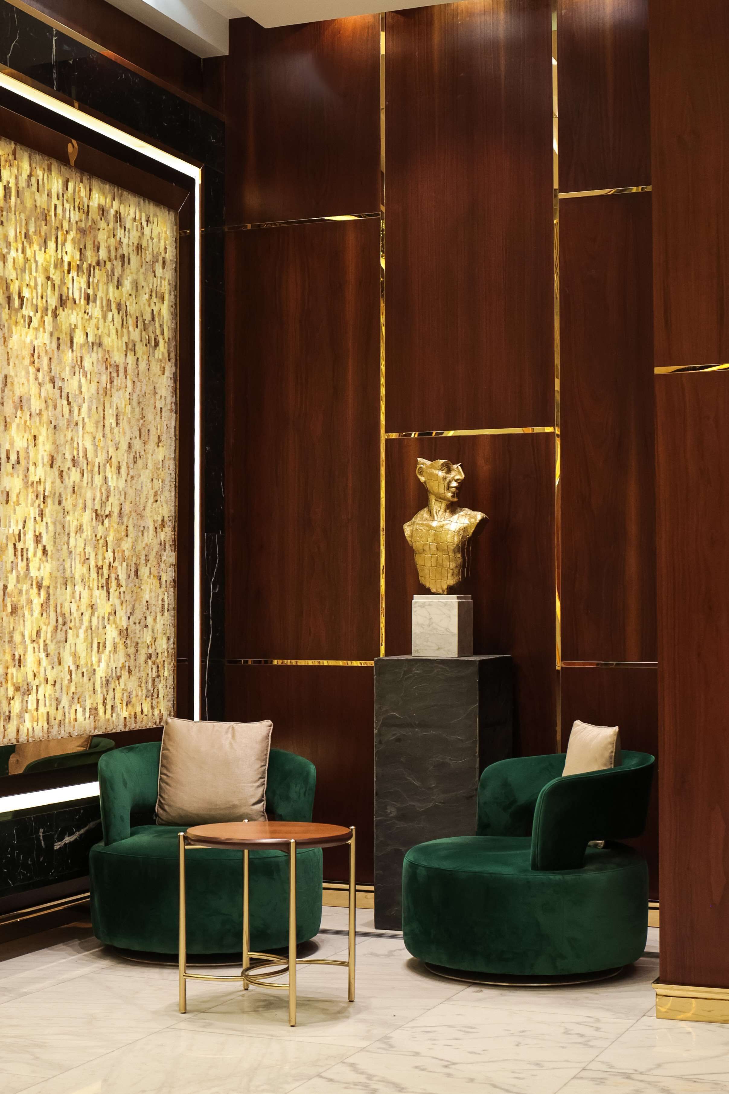 Un lobby lujoso con paneles de madera oscura, un sofá de terciopelo verde, una pequeña mesa redonda y una escultura de busto dorado.
