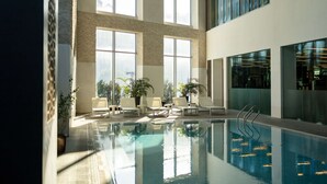 An indoor swimming pool with clear glass walls, large windows, and lounge chairs.