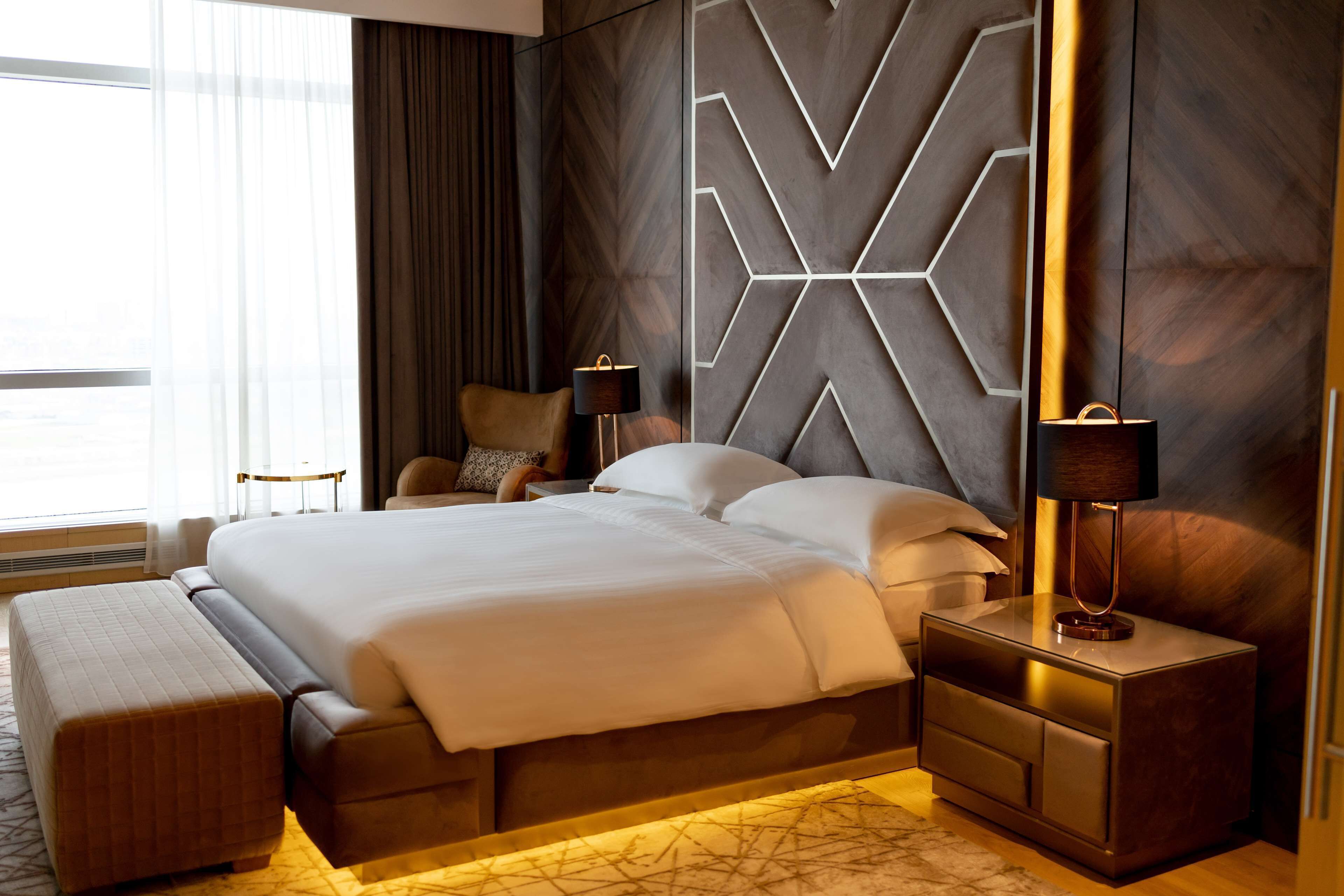 A modern hotel room with a large bed, a nightstand, a lamp, and a view of the city through the window.