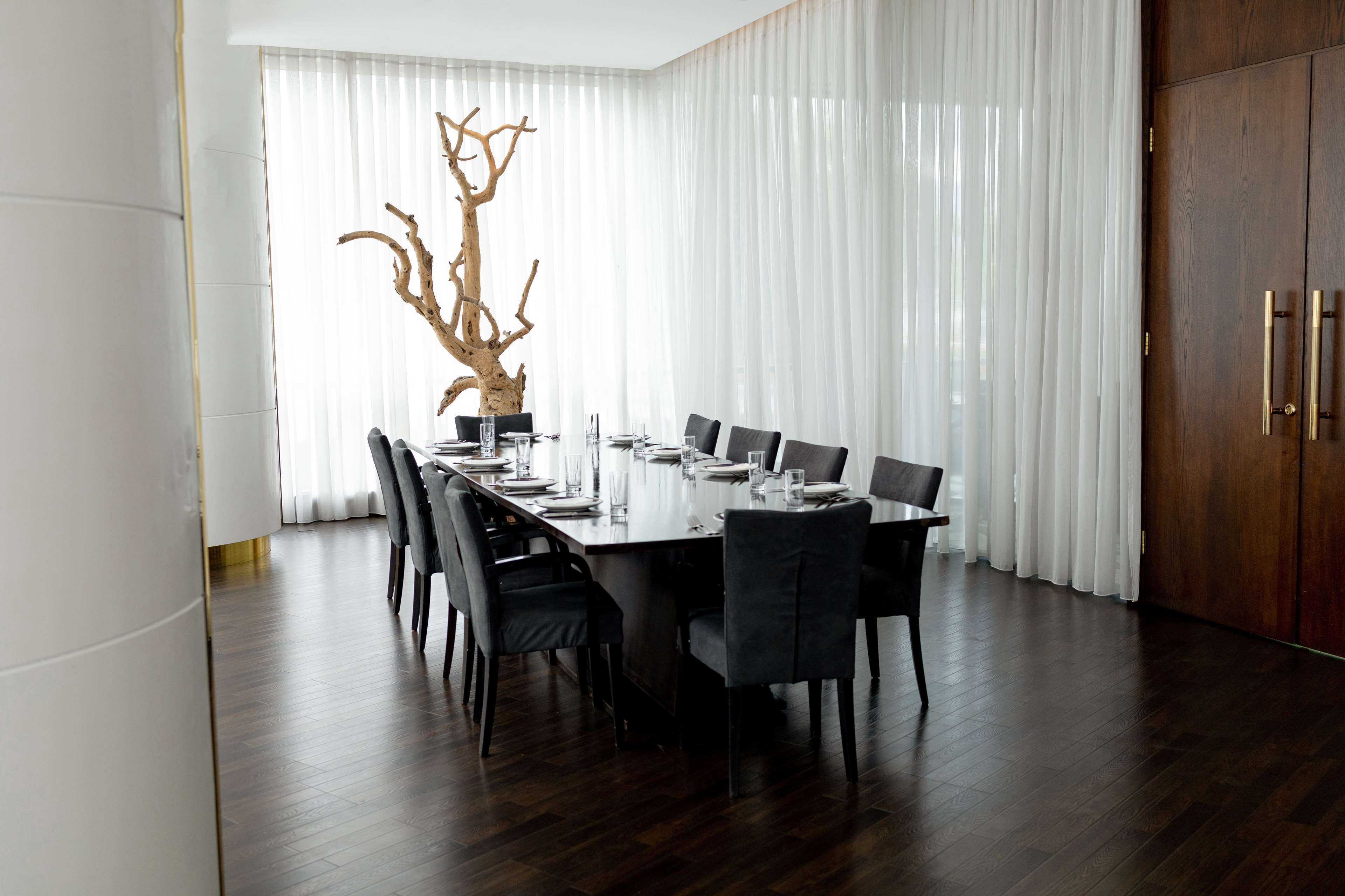 Une salle à manger moderne avec une longue table dressée pour six personnes, entourée de chaises aux couleurs sombres. Une grande sculpture d’arbre en bois naturel est placée sur la table. La pièce est dotée de rideaux blancs et d’un plancher en bois.