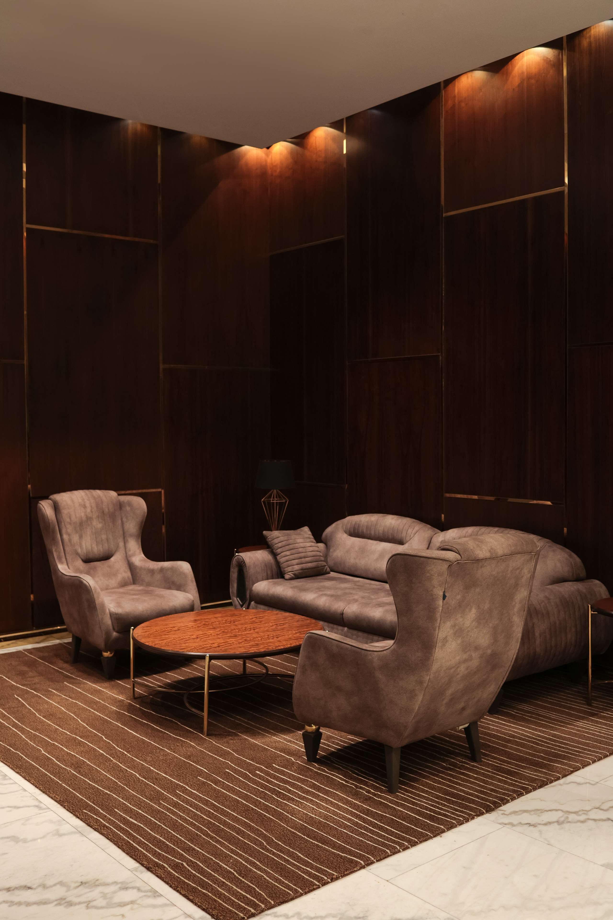 Ein modernes Wohnzimmer mit einem Ecksofa, einer Liege und einem kleinen runden Couchtisch. Der Raum verfügt über dunkelholzvertäfelte Wände und einen gemusterten Teppich auf dem Boden.