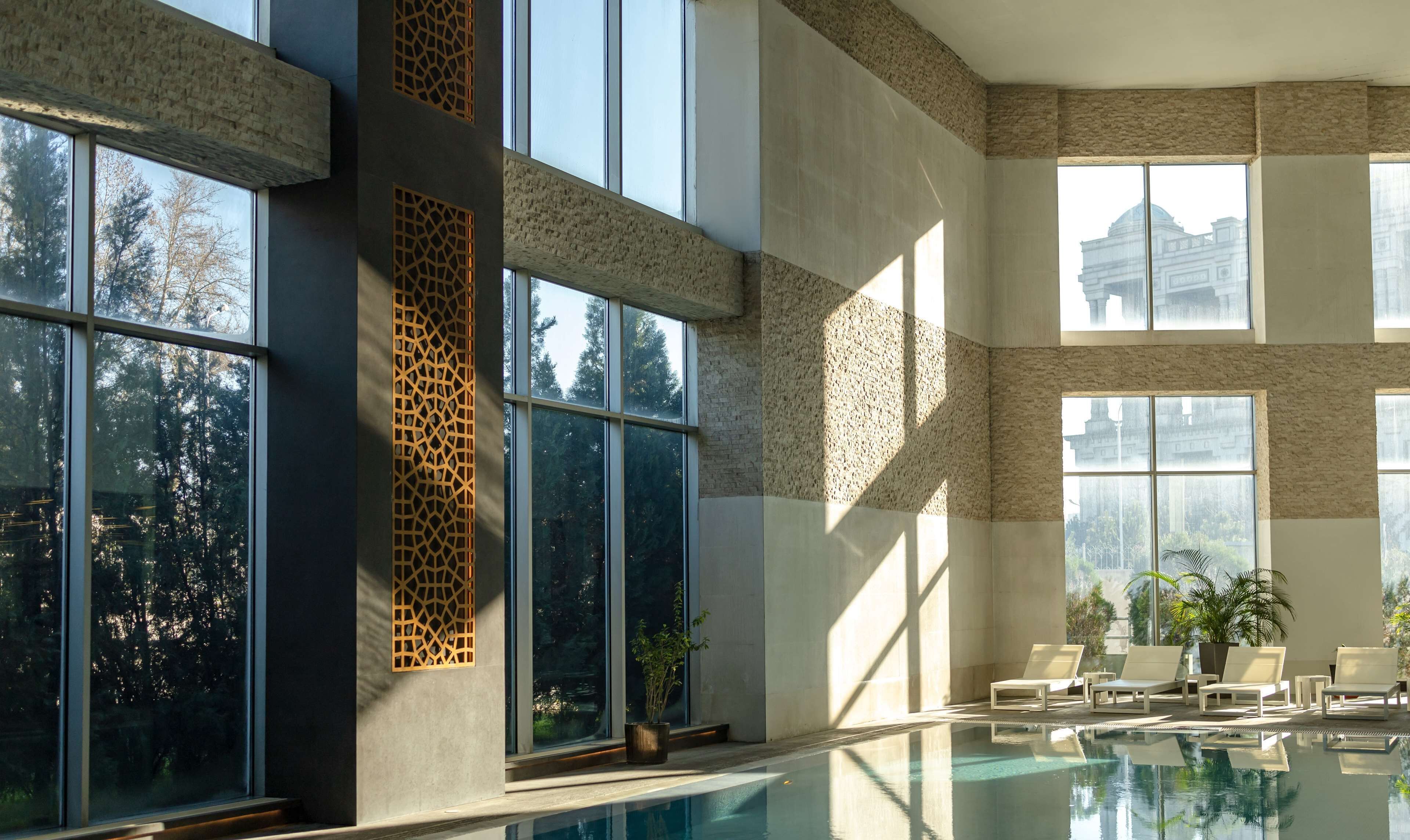A modern indoor pool area with large windows, a geometric wall design, and lounge chairs.