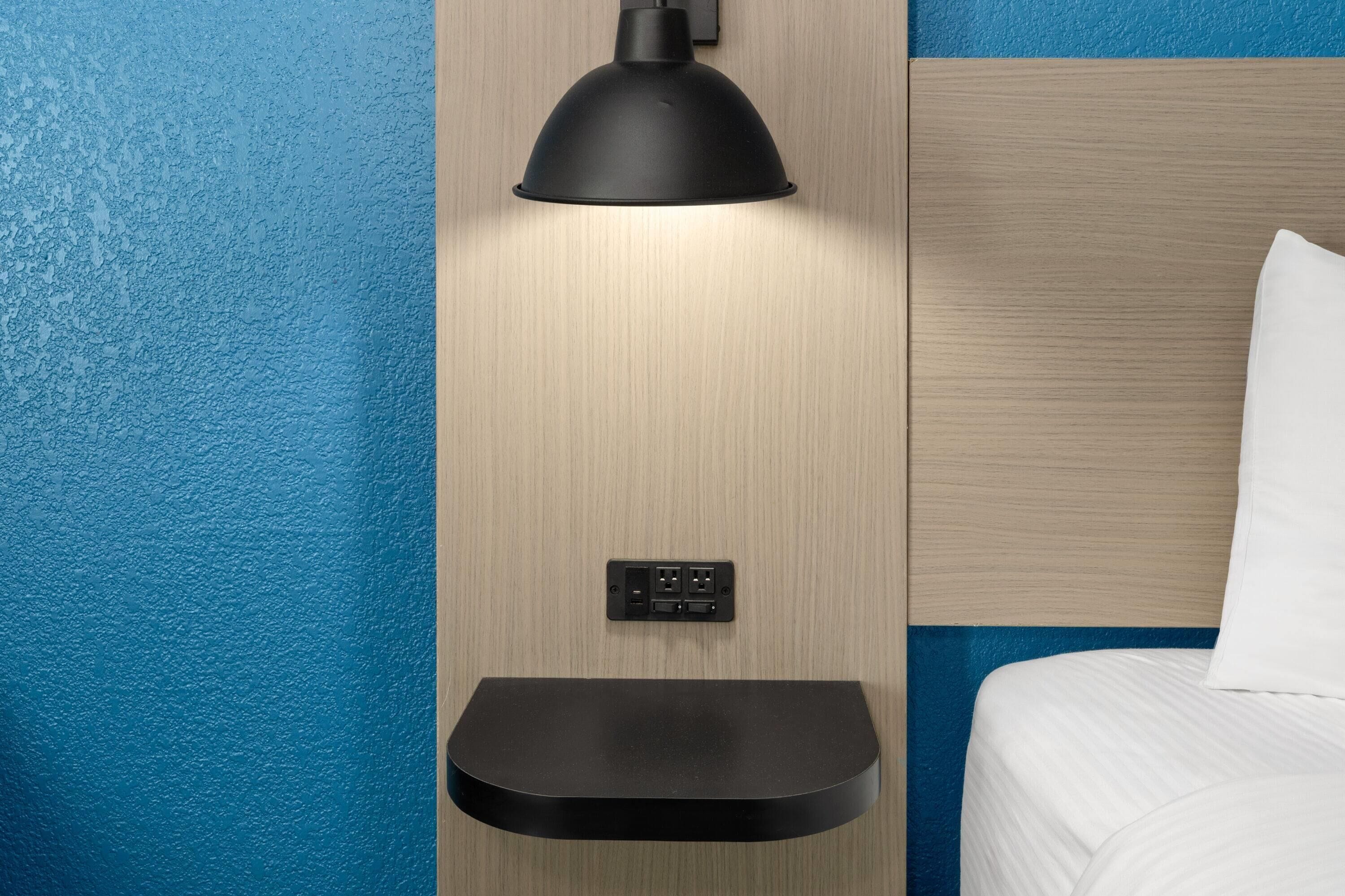 Ein modernes Hotelzimmer mit einem Holzkopfteil, einer schwarzen Wandlampe und einem kleinen Ablageschrank.