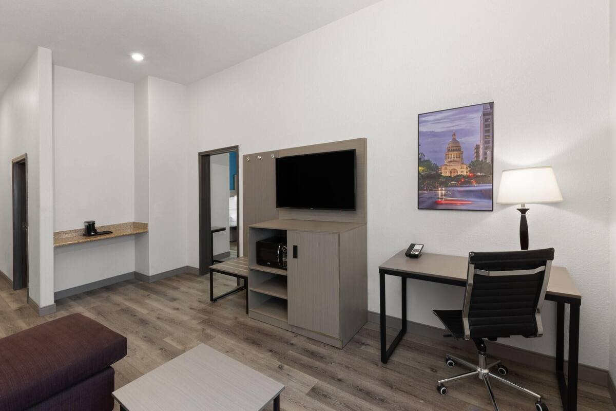 A hotel room with a desk, chair, TV, and a painting of a building.
