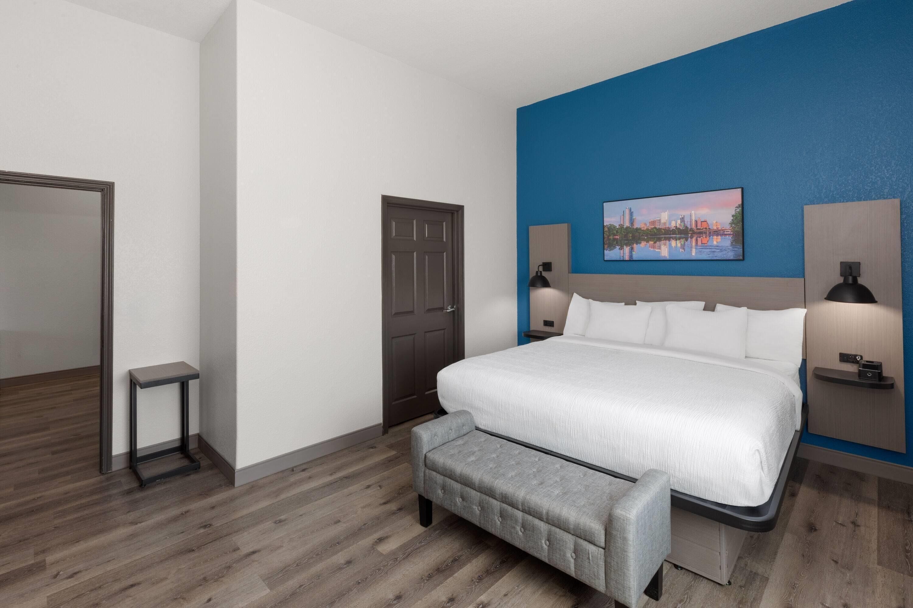 Moderni hotellihuone, jossa on suuri sänky, penkki, ovi ja seinään kiinnitetty valaisin.