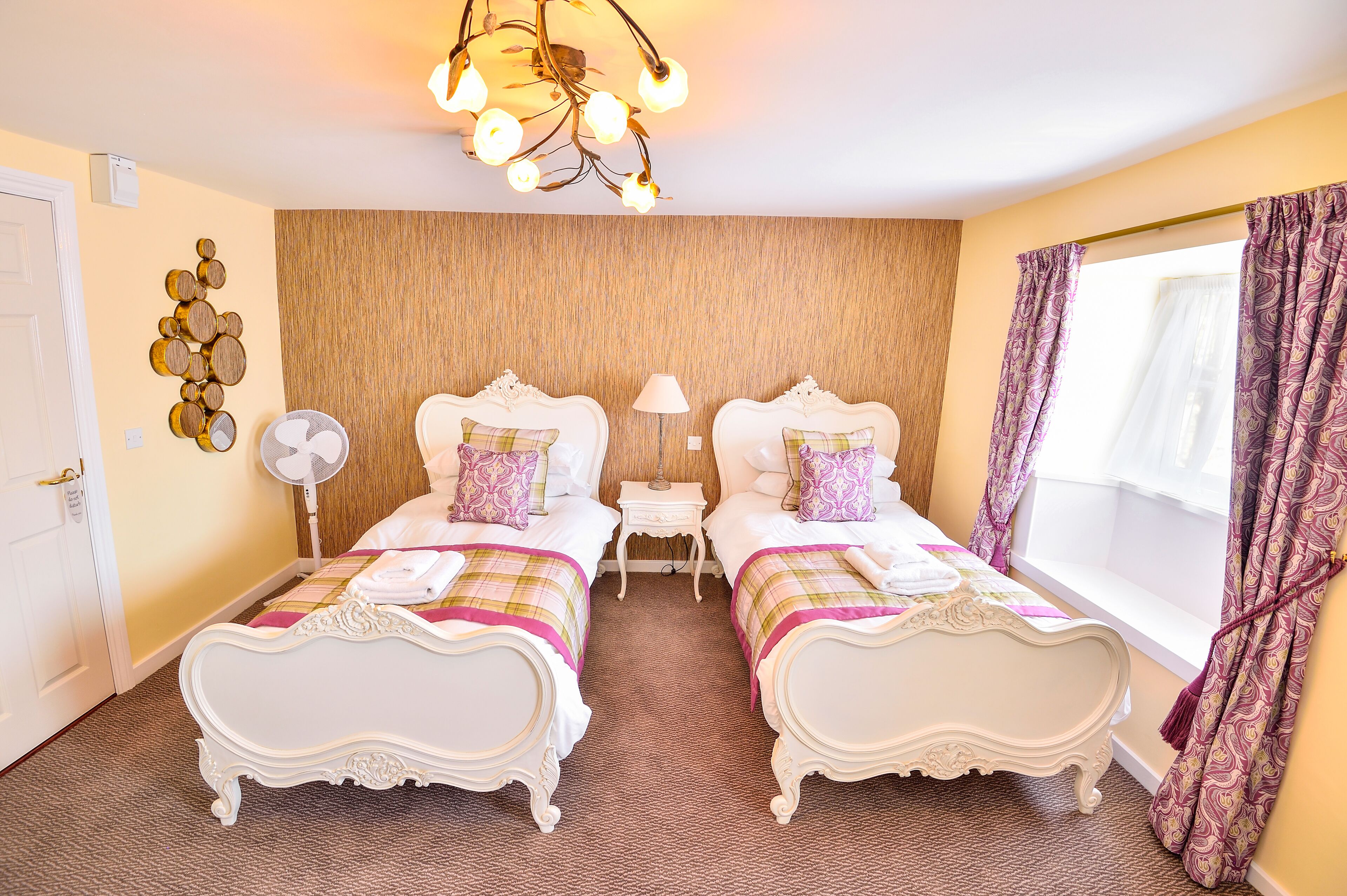 A hotel room with two ornate beds, a wooden accent wall, a chandelier, and a window with patterned curtains.