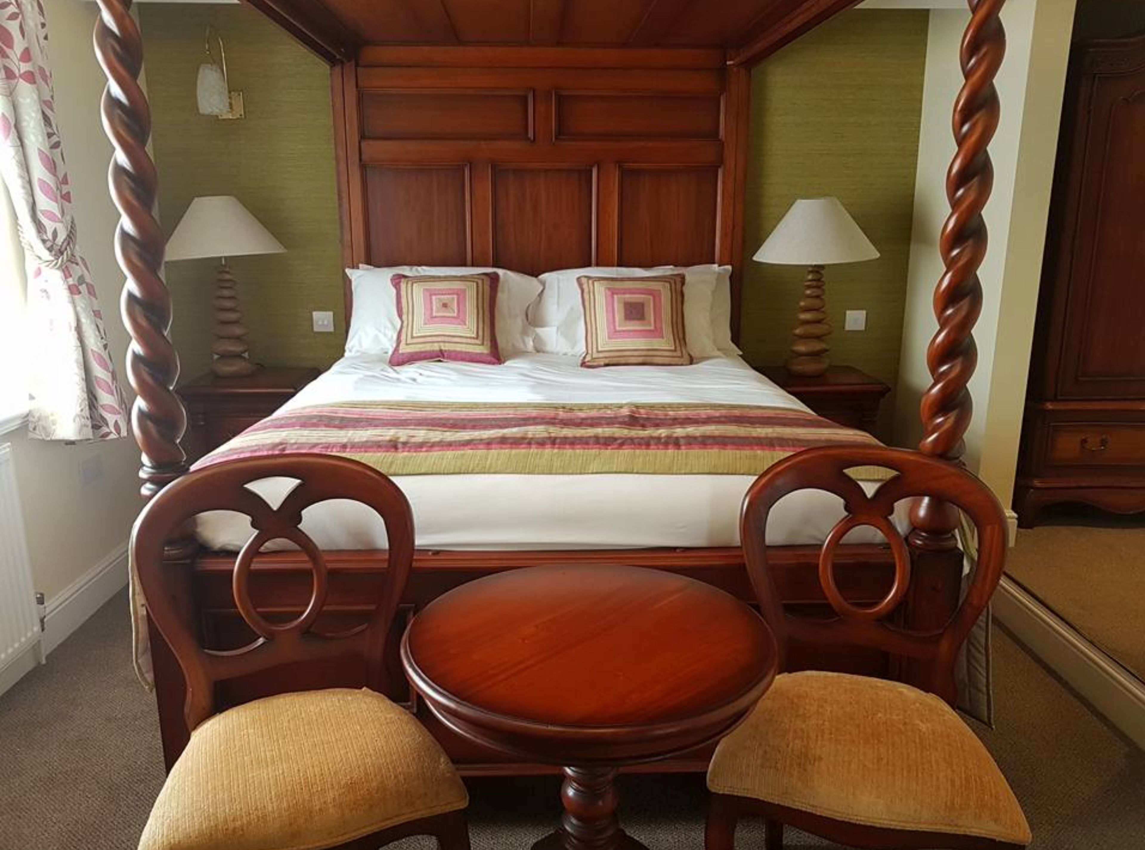 A four-poster bed with a wooden headboard, two bedside lamps, and a bedside table with a lamp. There are two upholstered chairs with cushions.