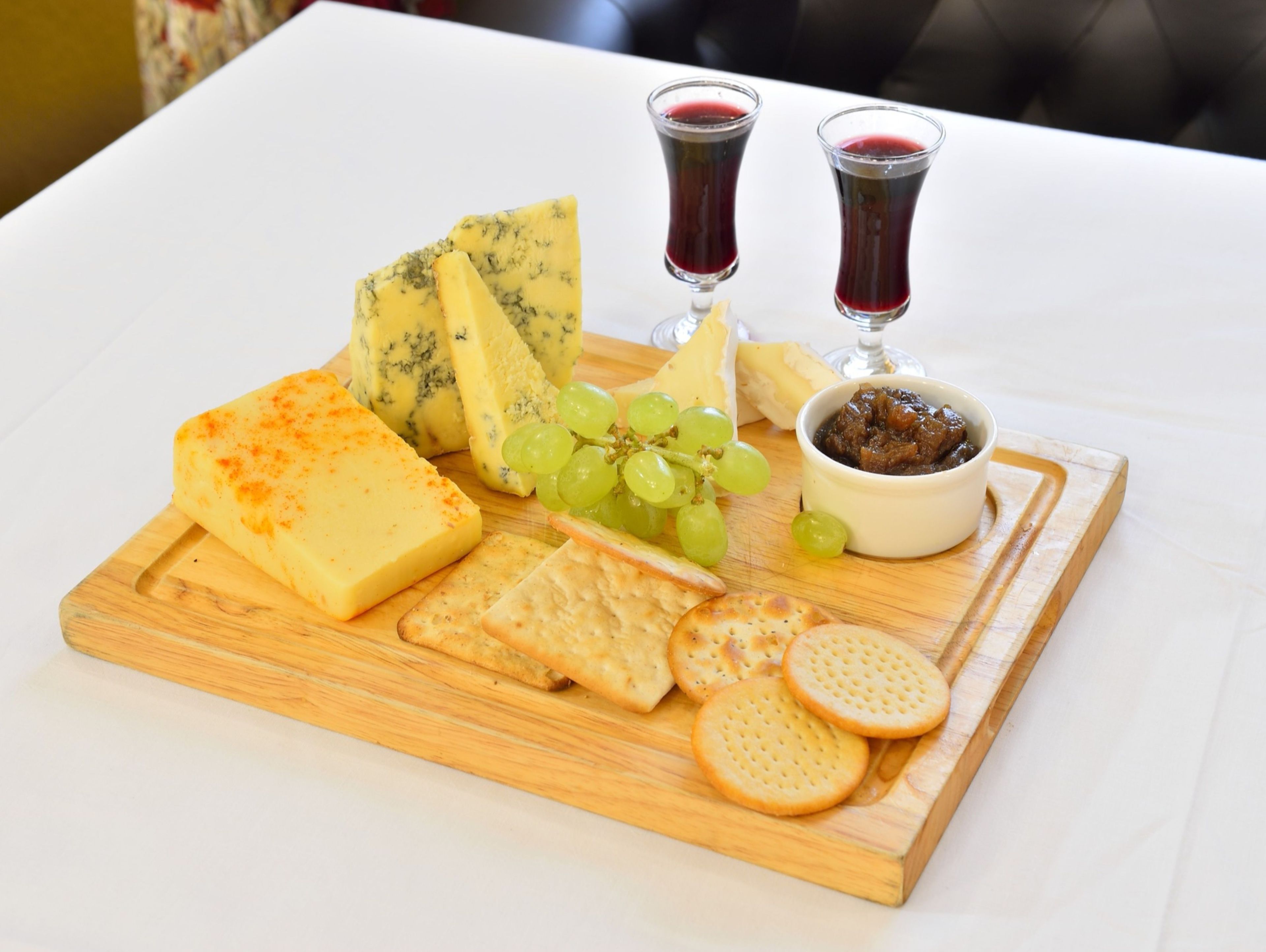 A wooden board with various cheeses, crackers, grapes, and two glasses of red wine.