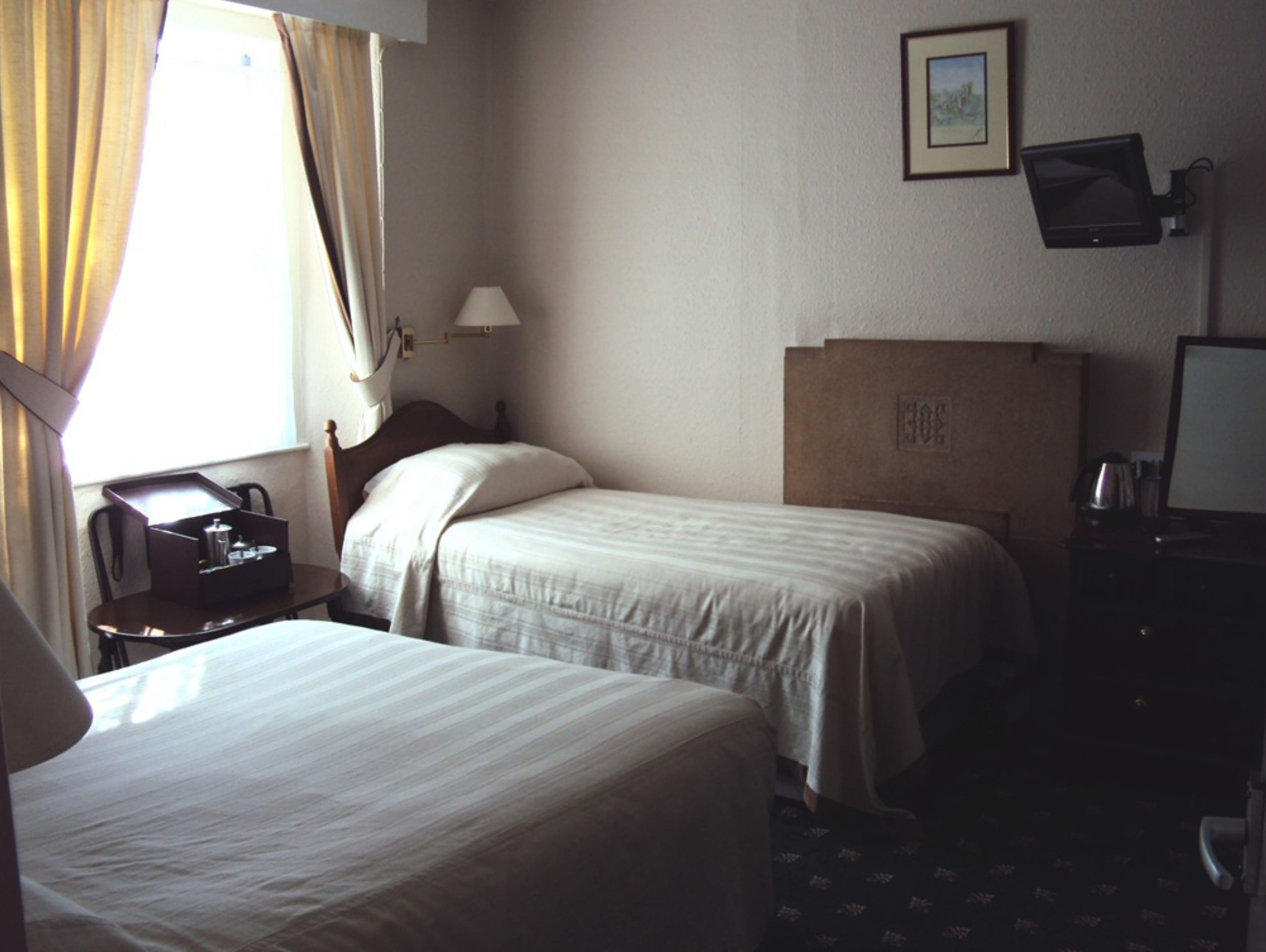 A hotel room with two single beds, a desk with a telephone, a lamp, and a television on the wall.