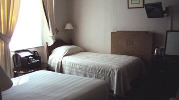 A hotel room with two single beds, a desk with a telephone, a lamp, and a television on the wall.