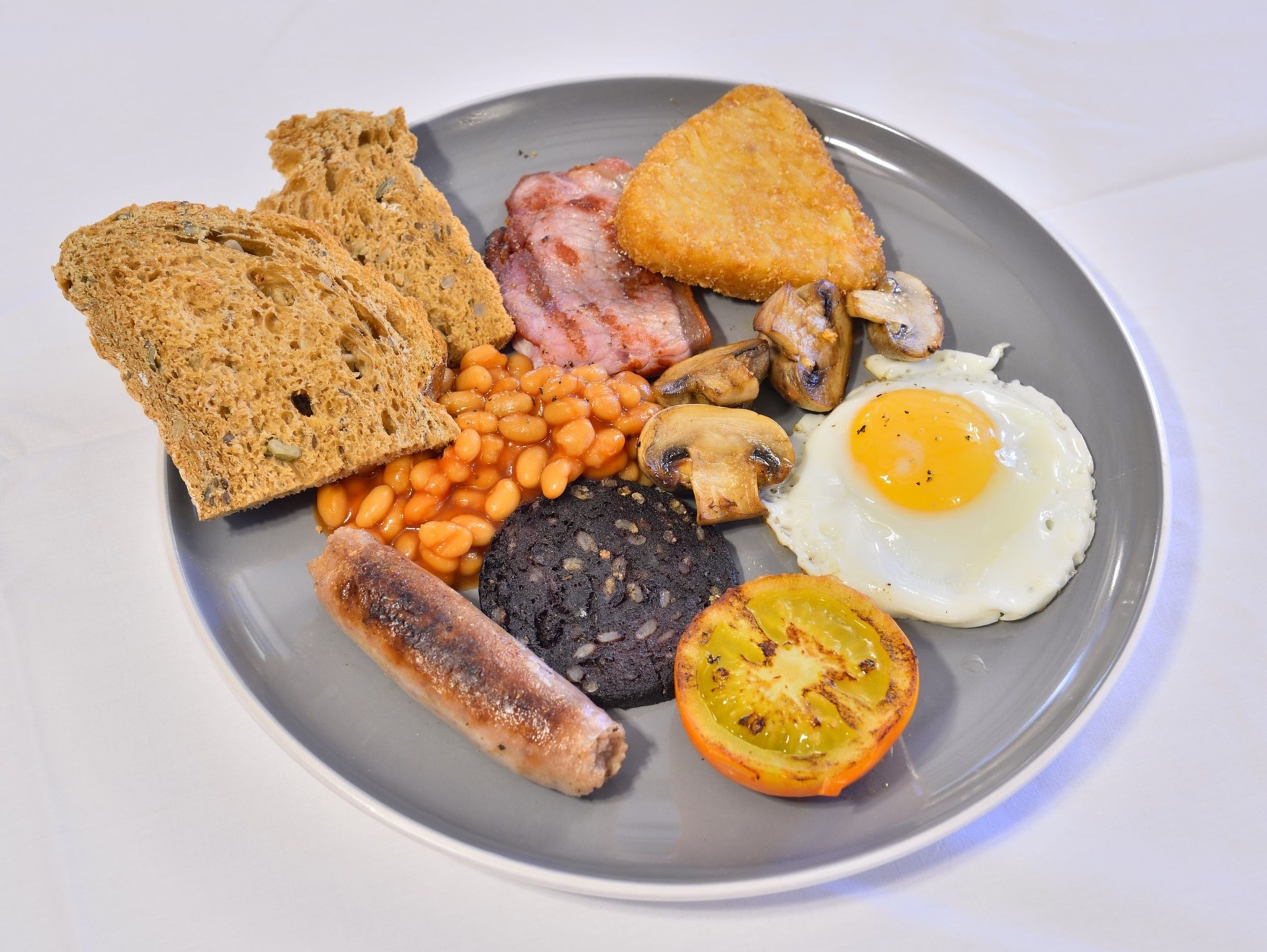 Een bord met toast, bonen, champignons, een gebakken ei, worst en gegrilde tomaat.