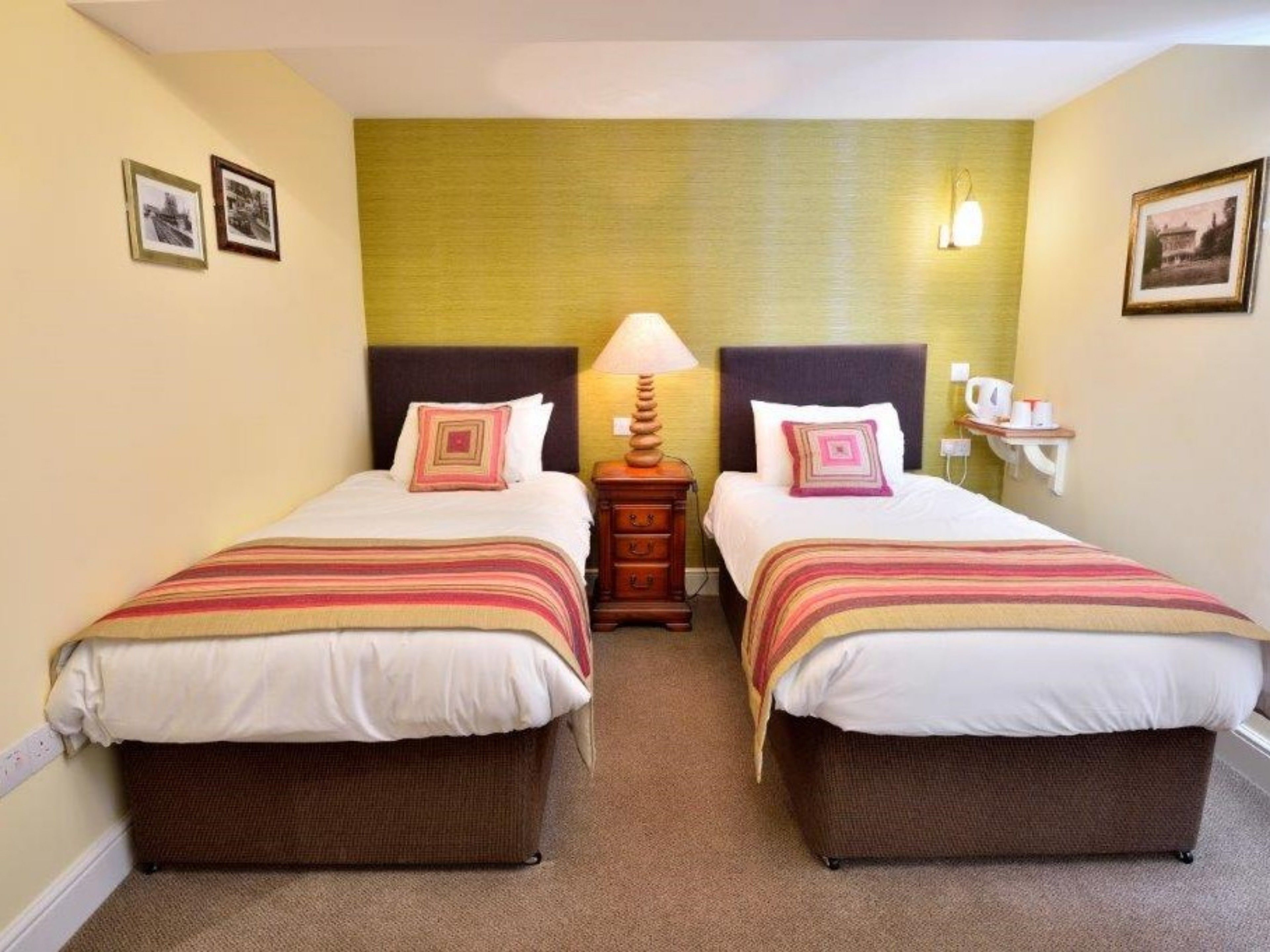 A hotel room with two beds, each with a striped bedspread and matching pillows. There is a nightstand between the beds with a lamp and a small table with a cup and saucer. The walls are painted in a light color, and there are framed pictures on the walls.