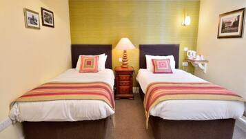 A hotel room with two beds, each with a striped bedspread and matching pillows. There is a nightstand between the beds with a lamp and a small table with a cup and saucer. The walls are painted in a light color, and there are framed pictures on the walls.