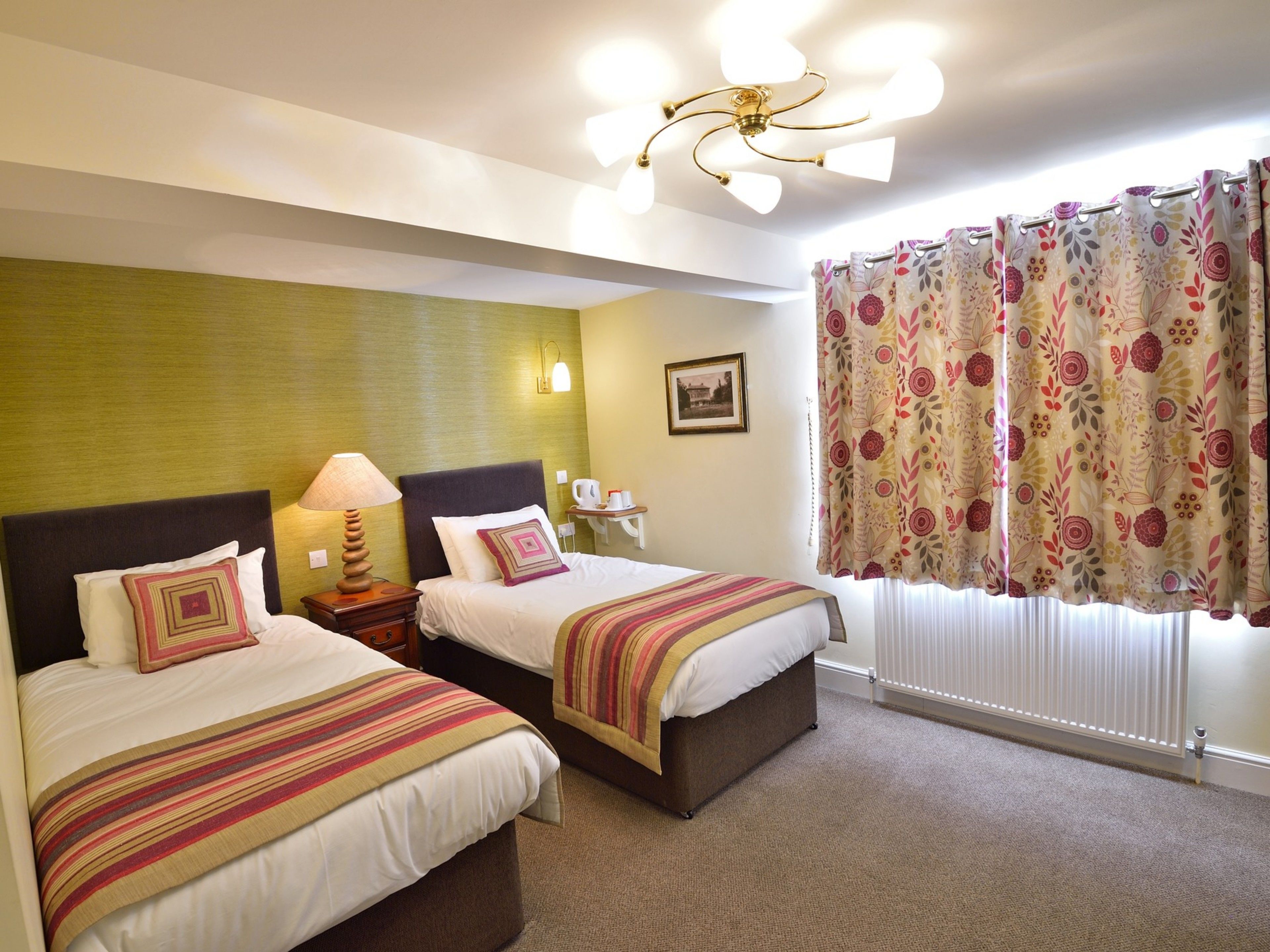 A hotel room with two beds, a ceiling fan, a window with floral curtains, and a framed picture on the wall.