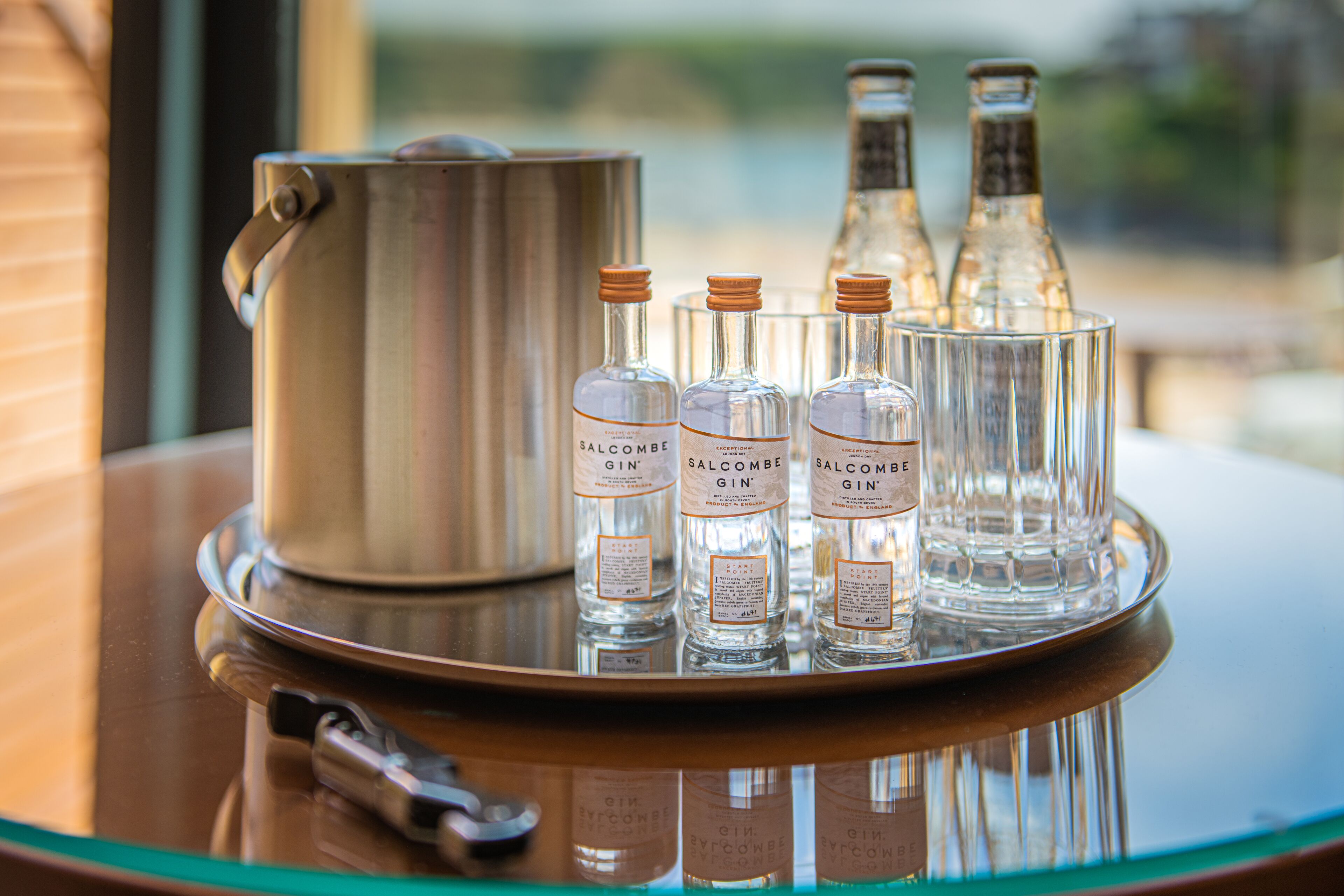 A tray with three bottles of Salcombe Gin and a metal pot on a reflective surface.