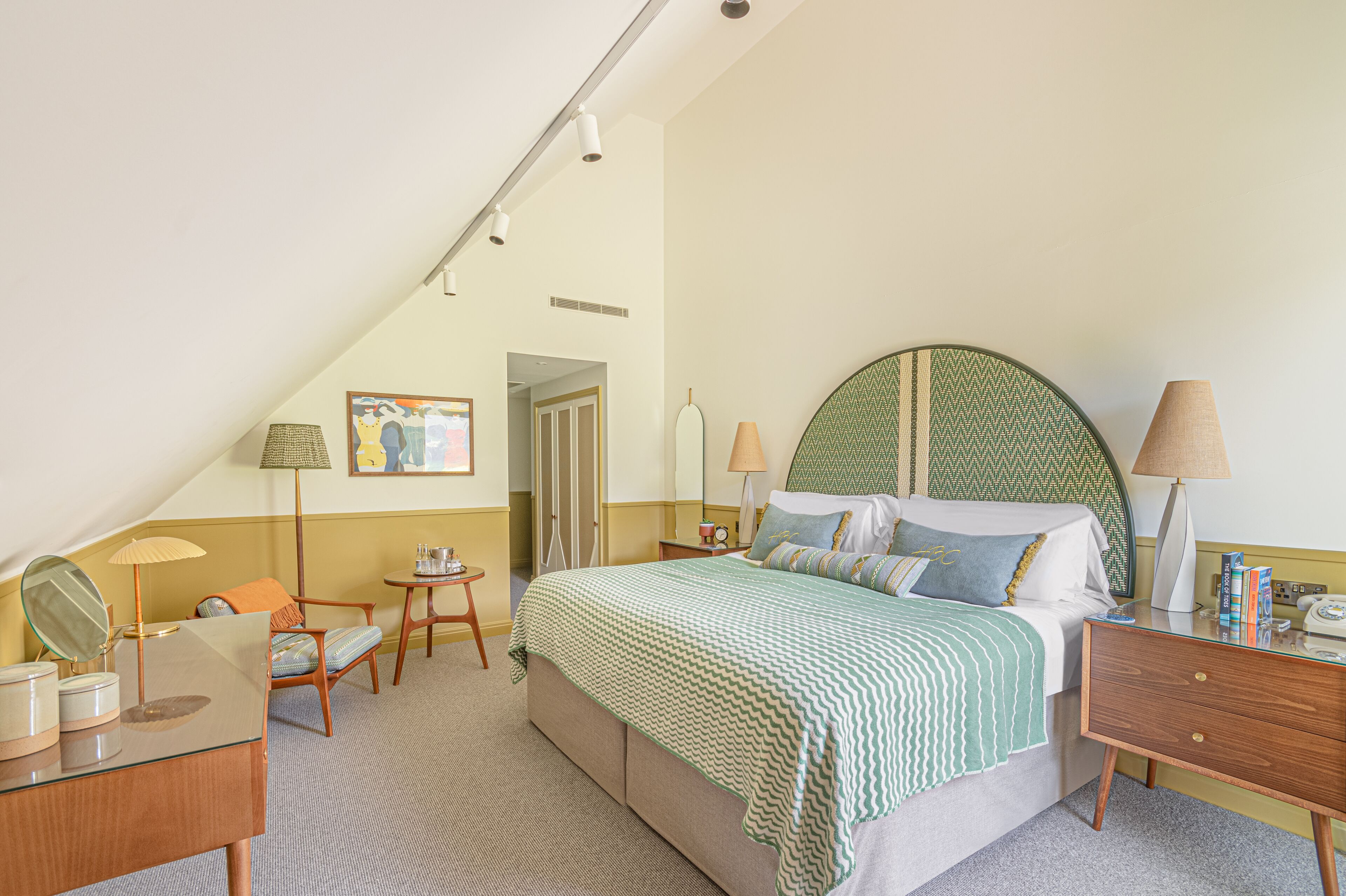A hotel room with a bed, bedside table, lamp, chair, and a small table with a lamp.