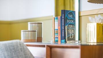 Habitación de hotel con un escritorio de madera, una silla, una mesita de noche con una caja de pañuelos y una estantería con libros.