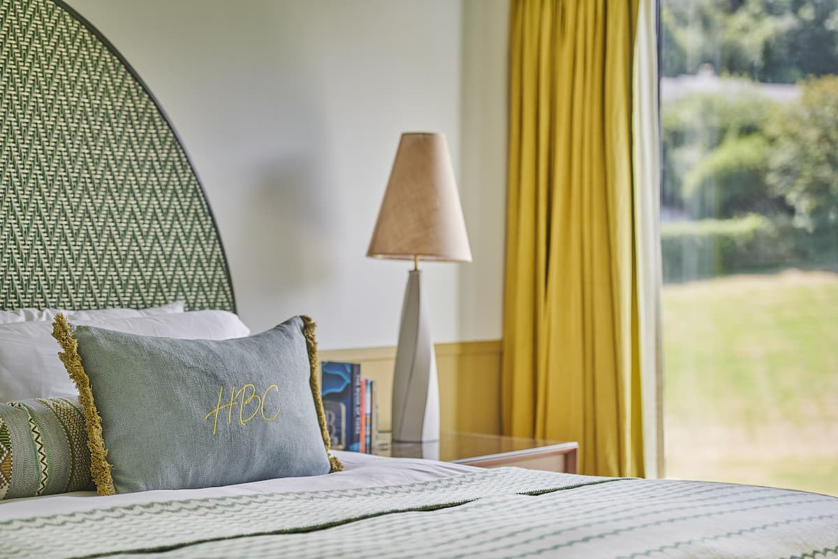 A neatly made bed with patterned bedding and pillows, a bedside lamp, and a view of greenery through a window.