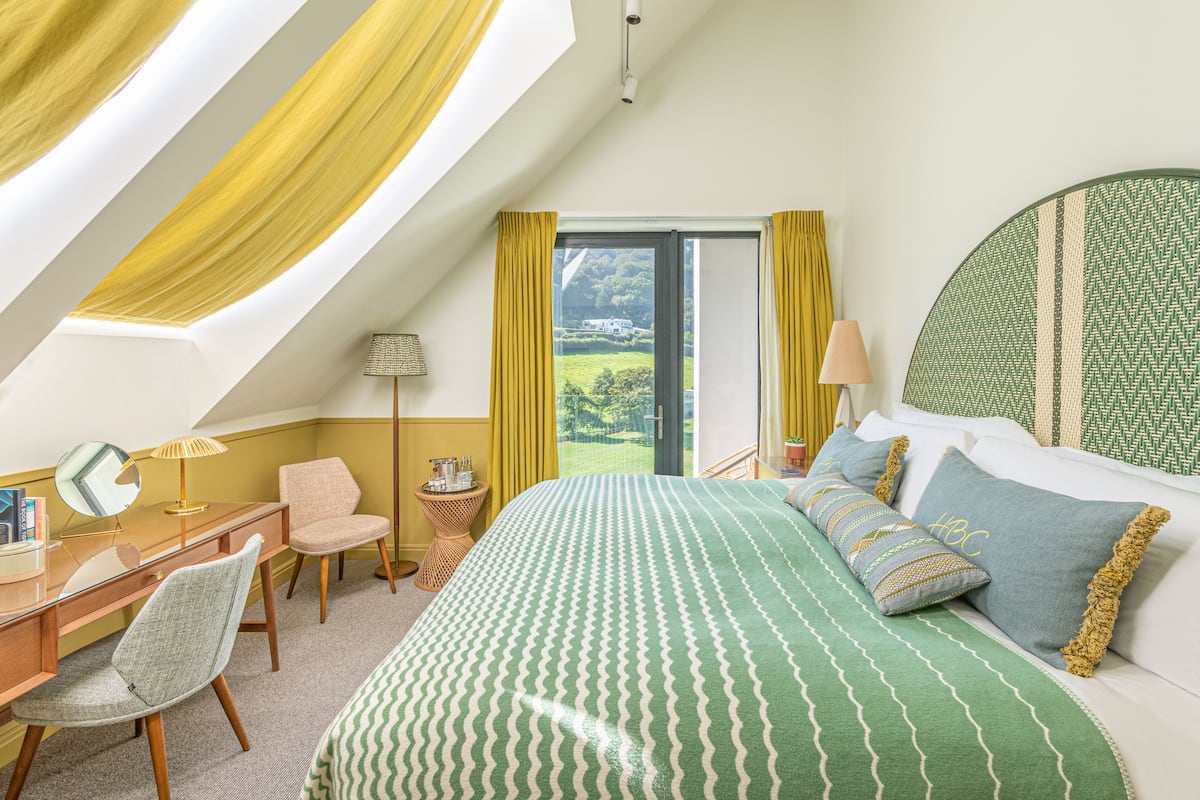 A bedroom with a large bed, a desk, a chair, and a view of a green landscape through a window.