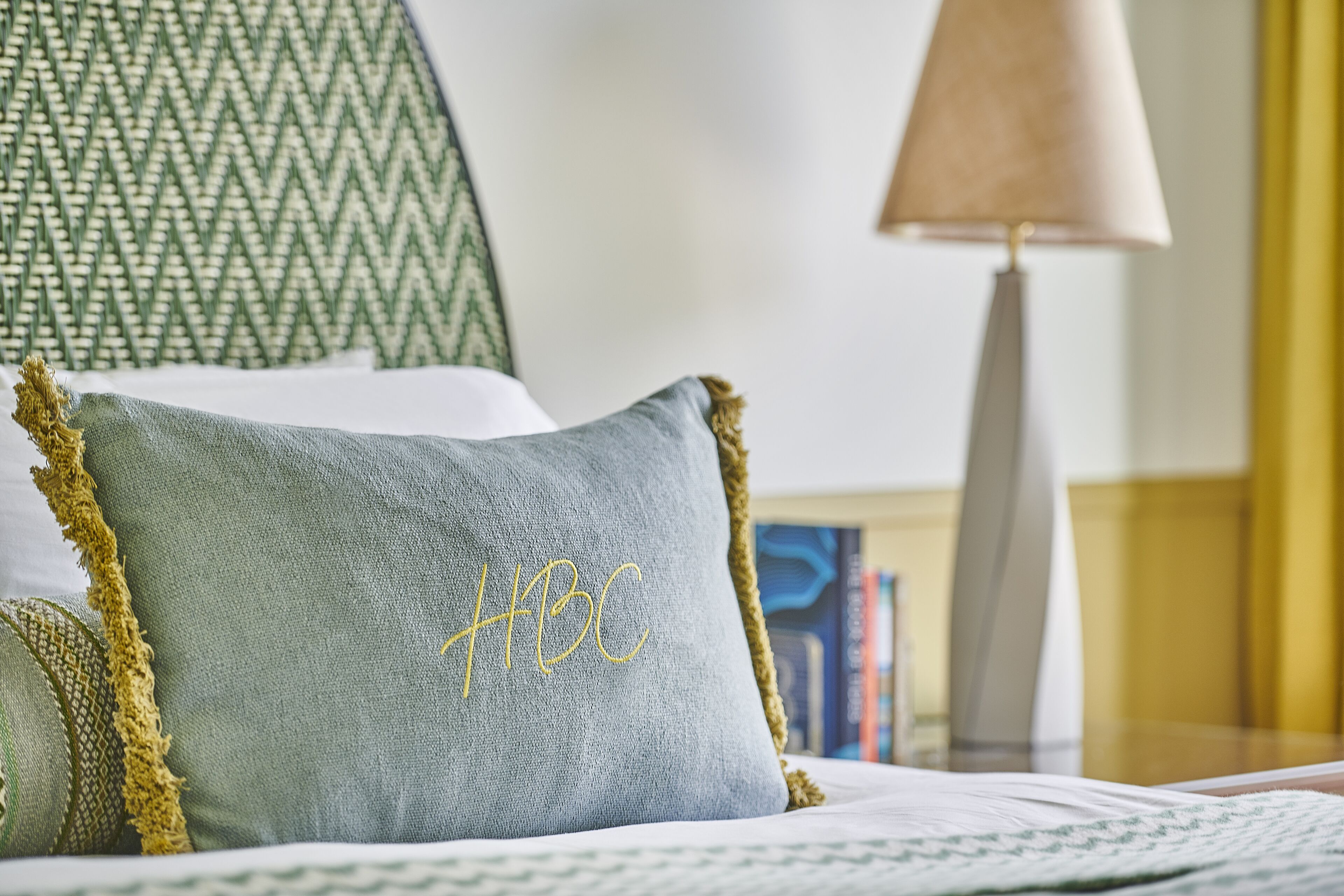 A bed with a green pillow embroidered with "HBC" and a lamp in the background.