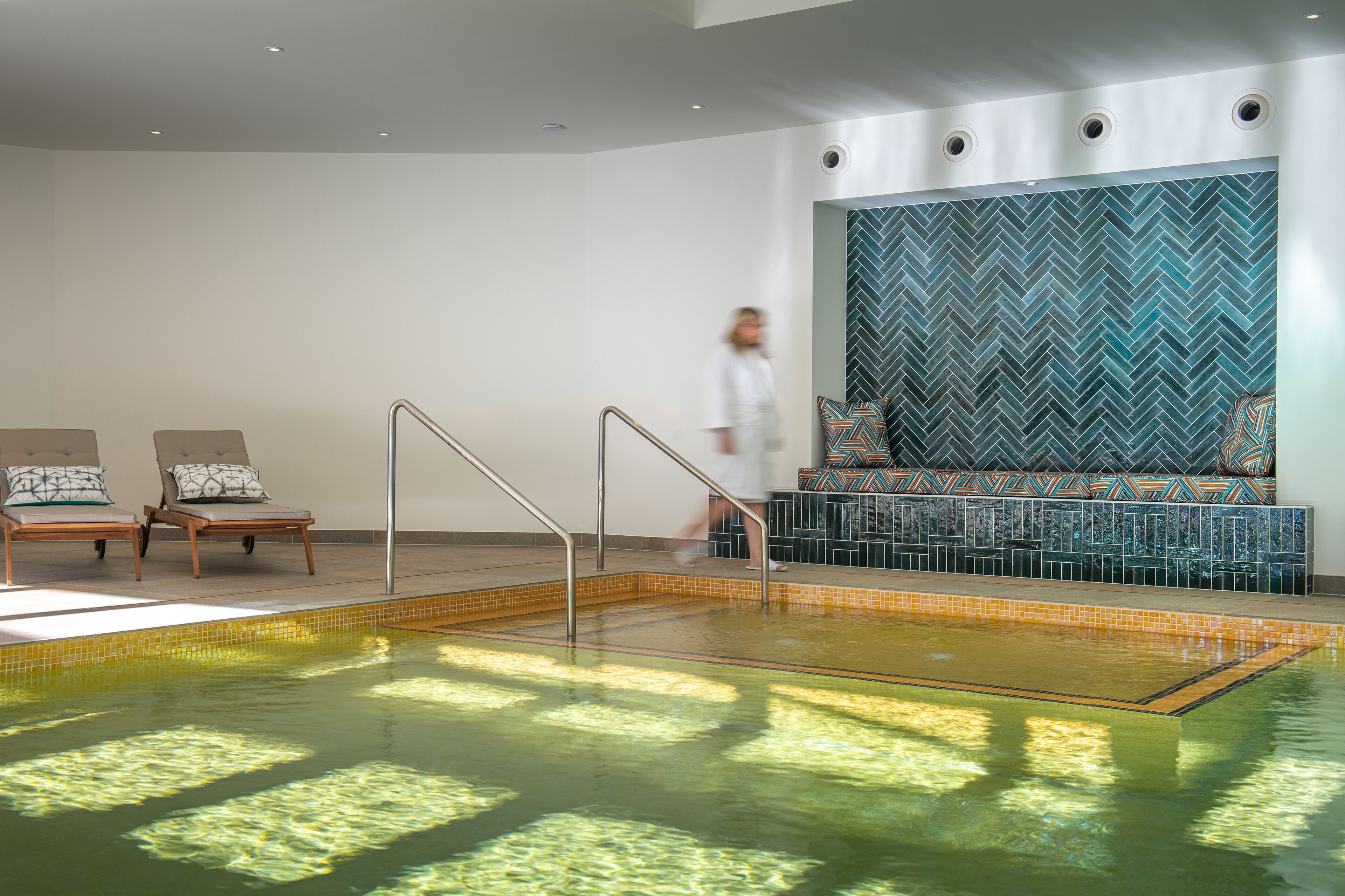 A modern indoor swimming pool with a herringbone tile wall, two lounge chairs, and a staircase leading to a seating area.