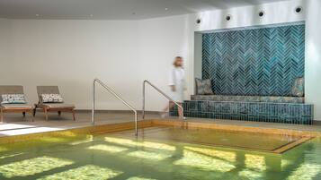 A modern indoor swimming pool with a herringbone tile wall, two lounge chairs, and a staircase leading to a seating area.